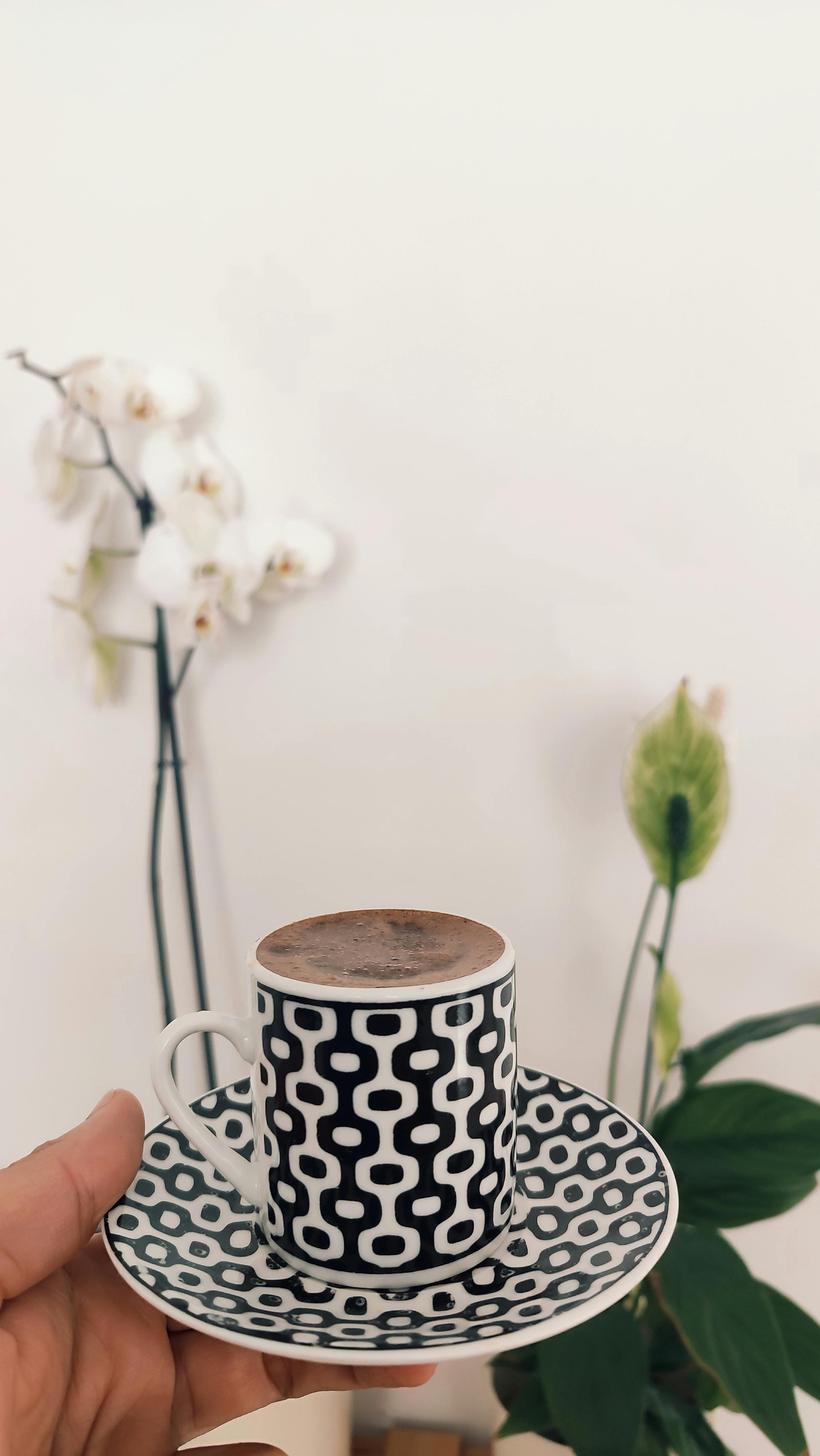 ฟรี ถ้วยกาแฟสีดำขาวดีไซน์เก๋ ลวดลายเรขาคณิต ถืออยู่ในห้องที่มีดอกกล้วยไม้สีขาวเป็นฉากหลัง คลังภาพถ่าย