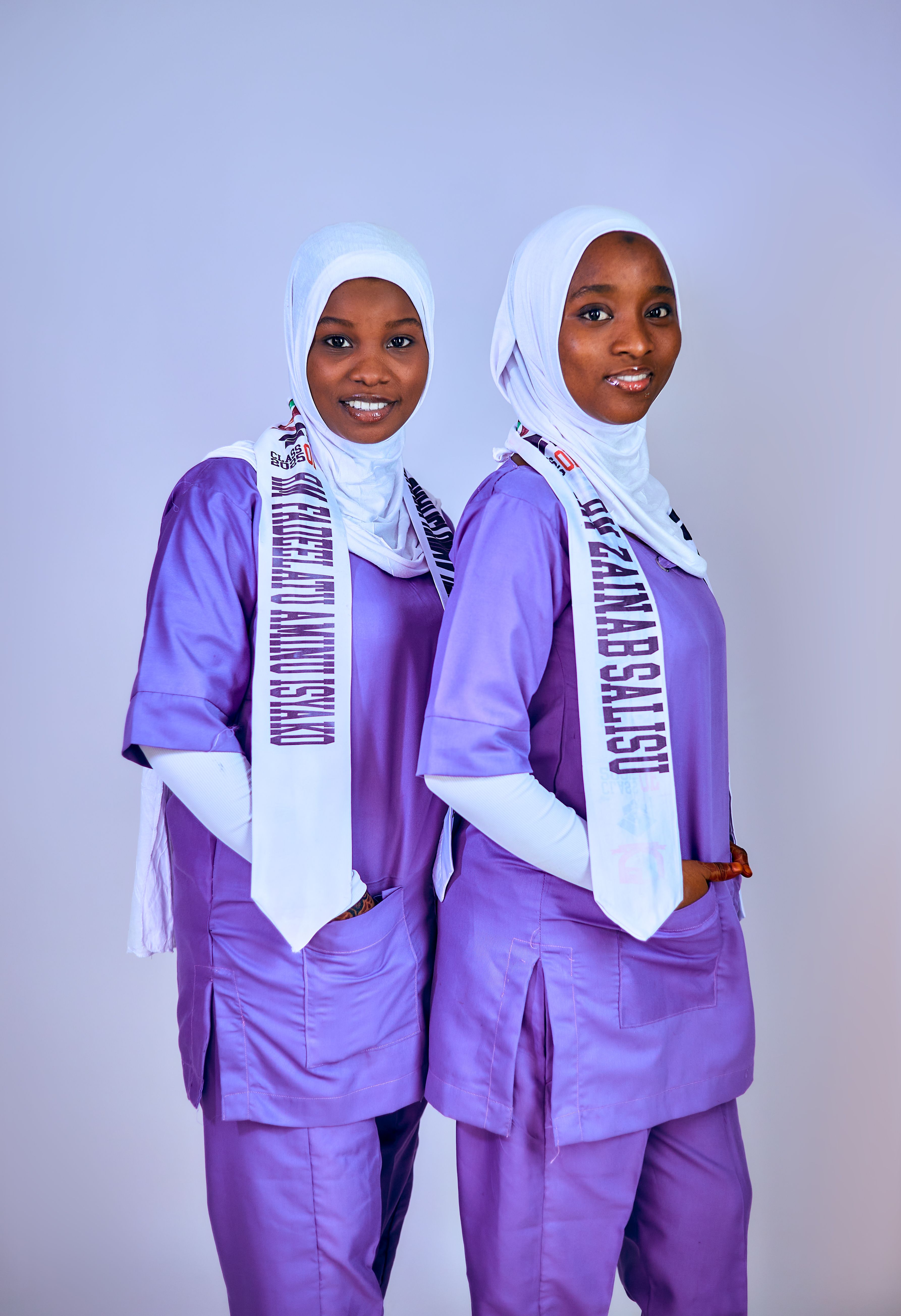 Two women wearing purple traditional attire with white hijabs pose against a plain background.
