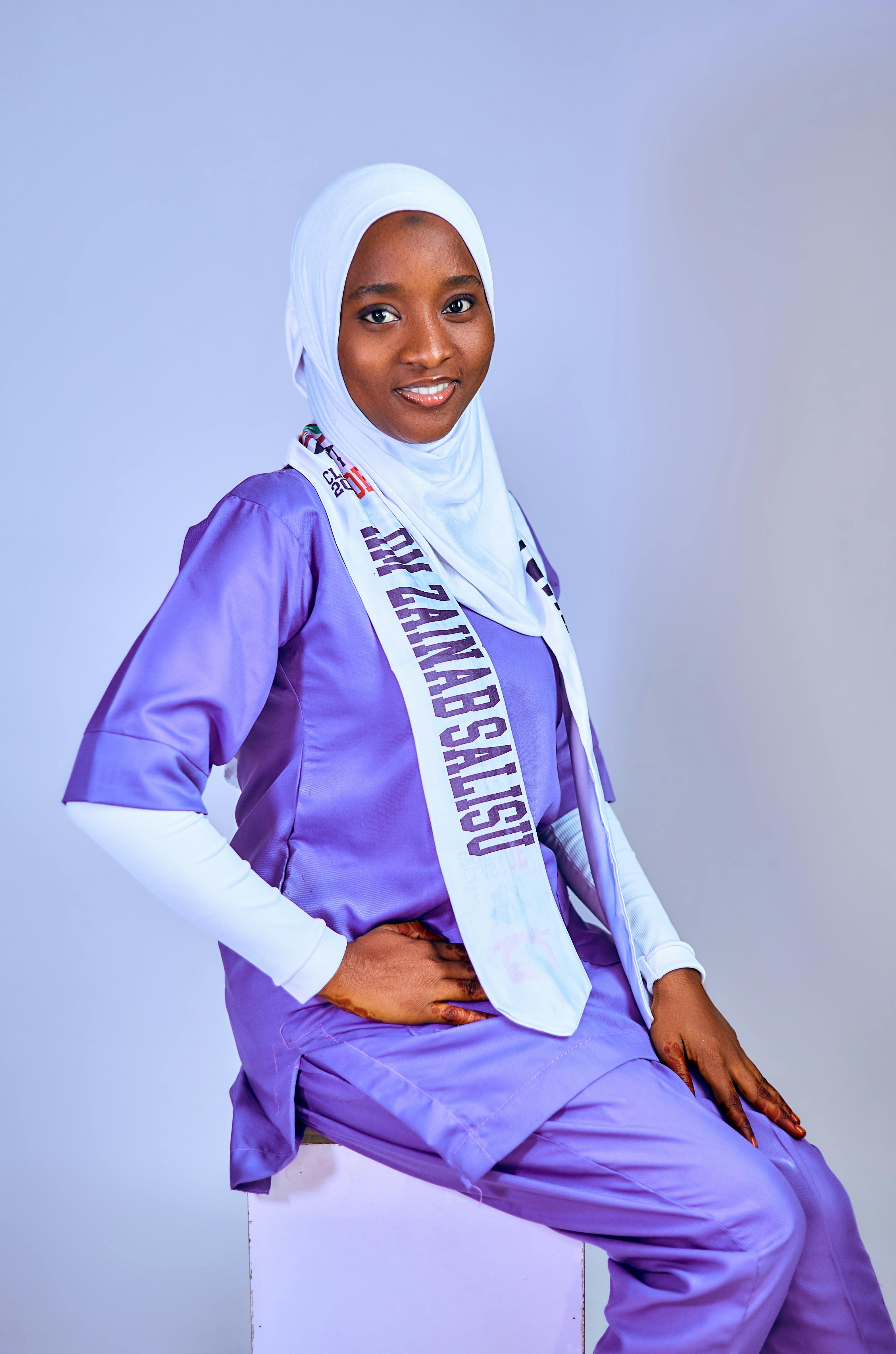 Portrait of a smiling woman in purple attire wearing a white hijab and a sash, sitting indoors.