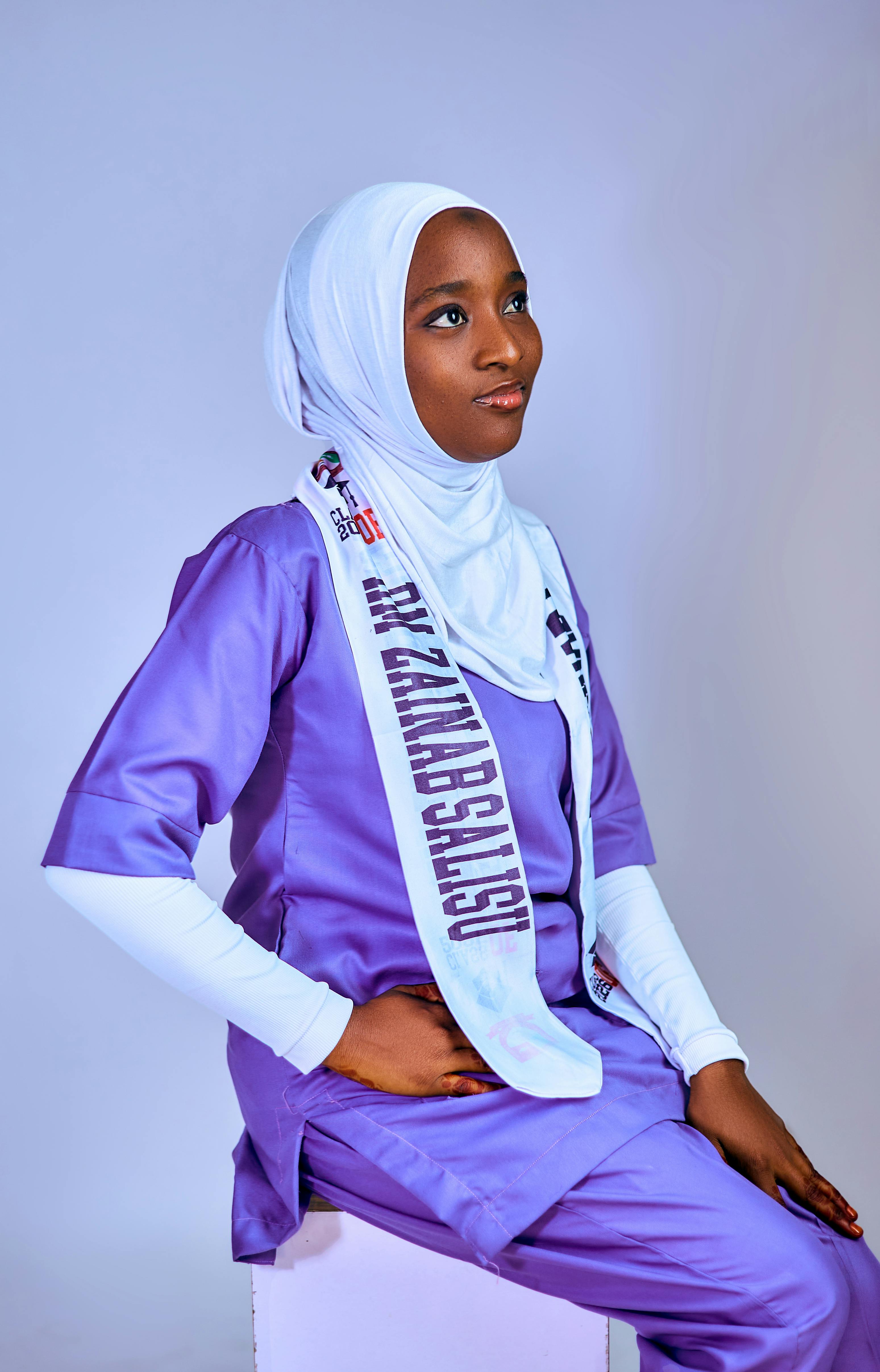 Young woman wearing hijab and purple scrubs, sitting confidently with a sash.