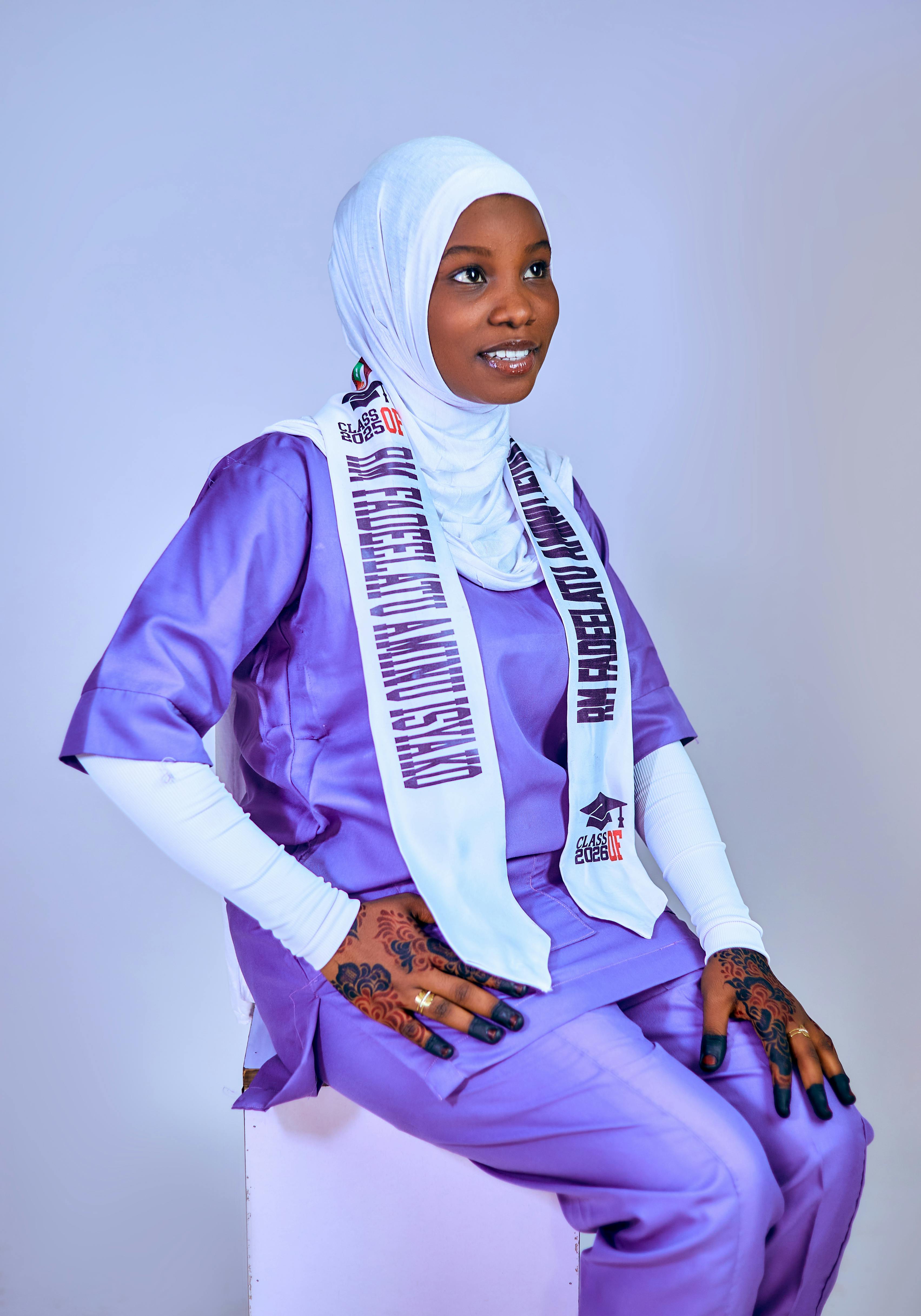 Young woman in purple graduation attire, seated, smiling at ceremony.