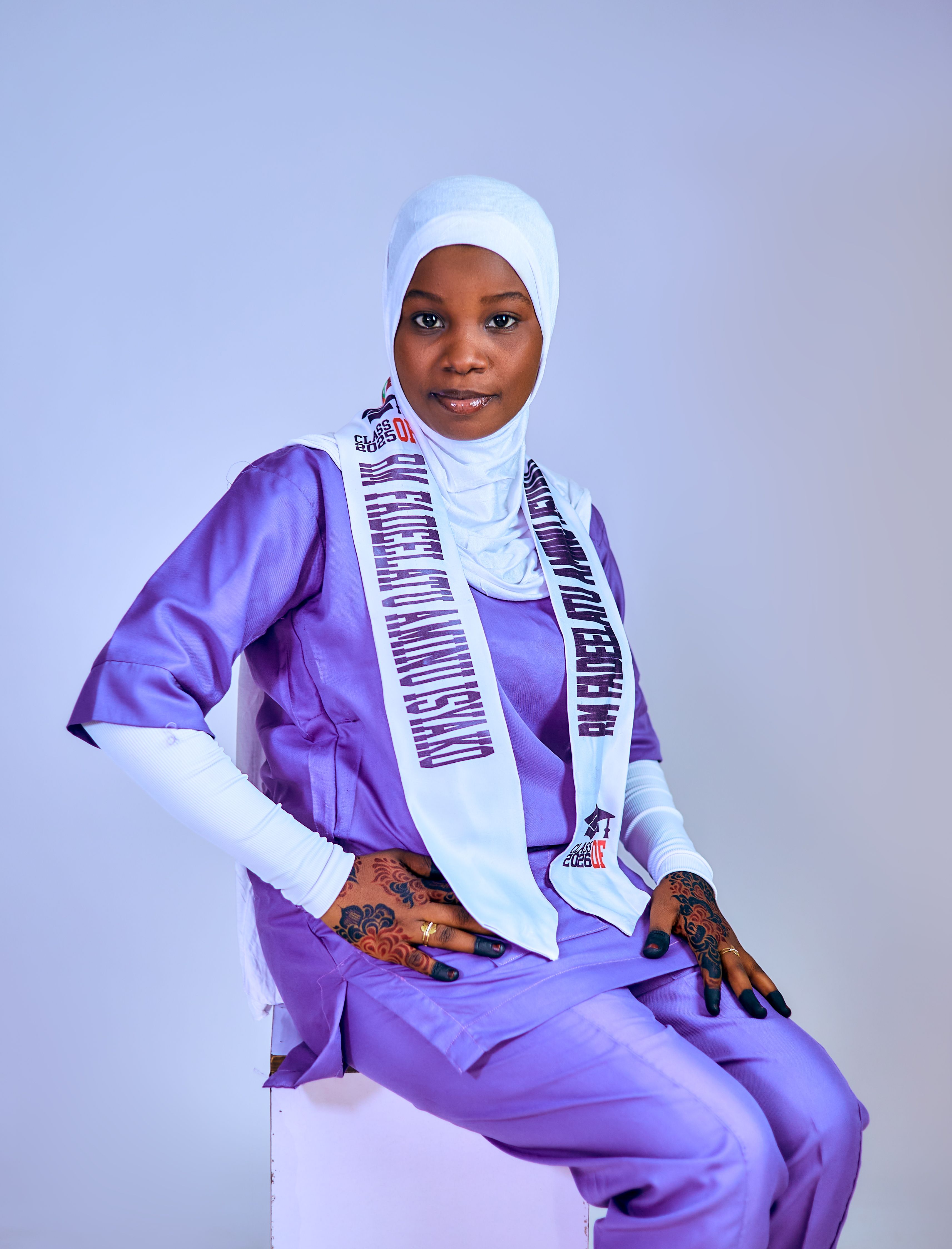 African woman in purple outfit and hijab with henna tattoo, seated elegantly.