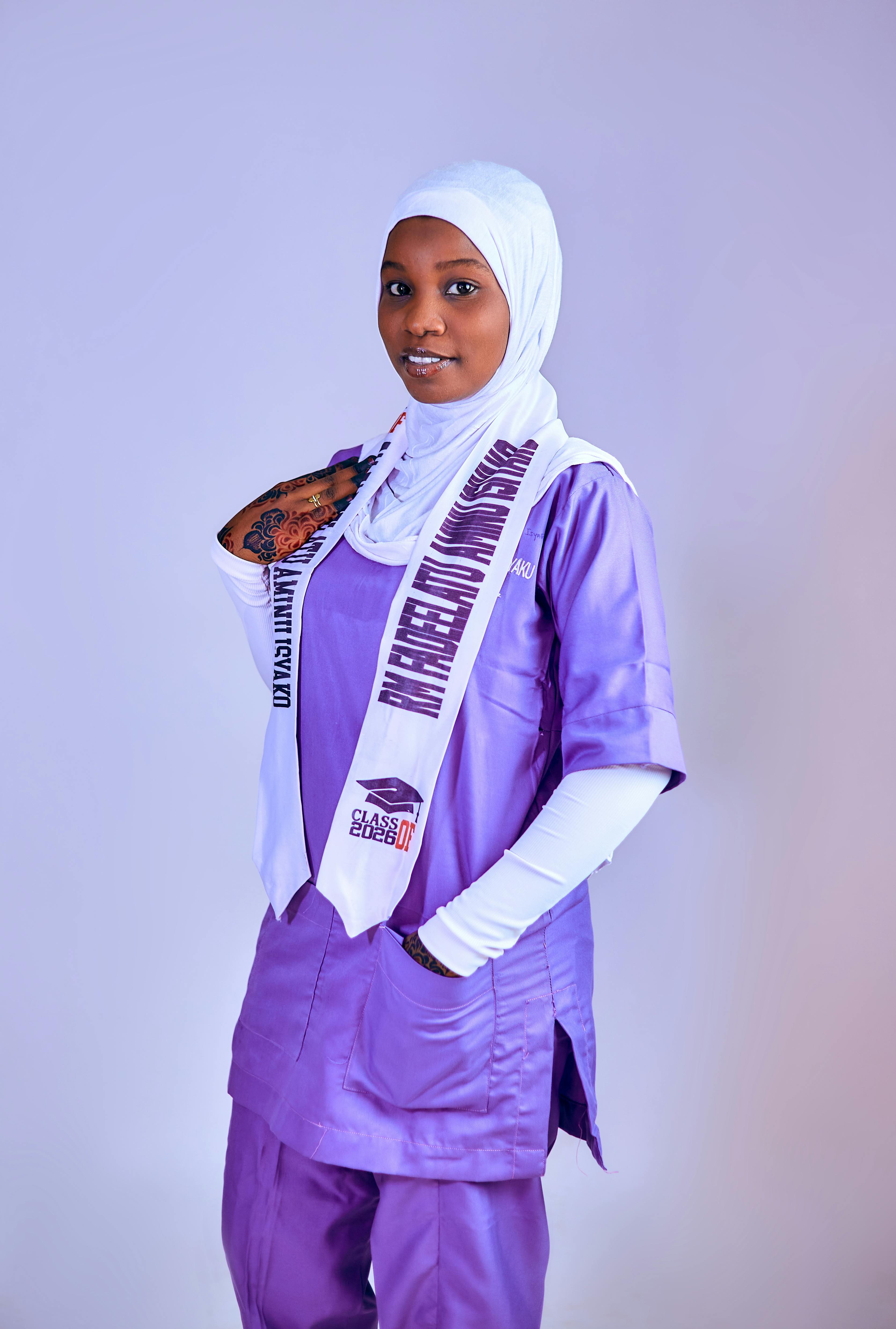 Confident nurse in purple scrubs and hijab, wearing a celebratory sash, exuding positivity.