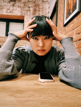 Person in cozy sweater looking contemplative at a table with a smartphone indoors.