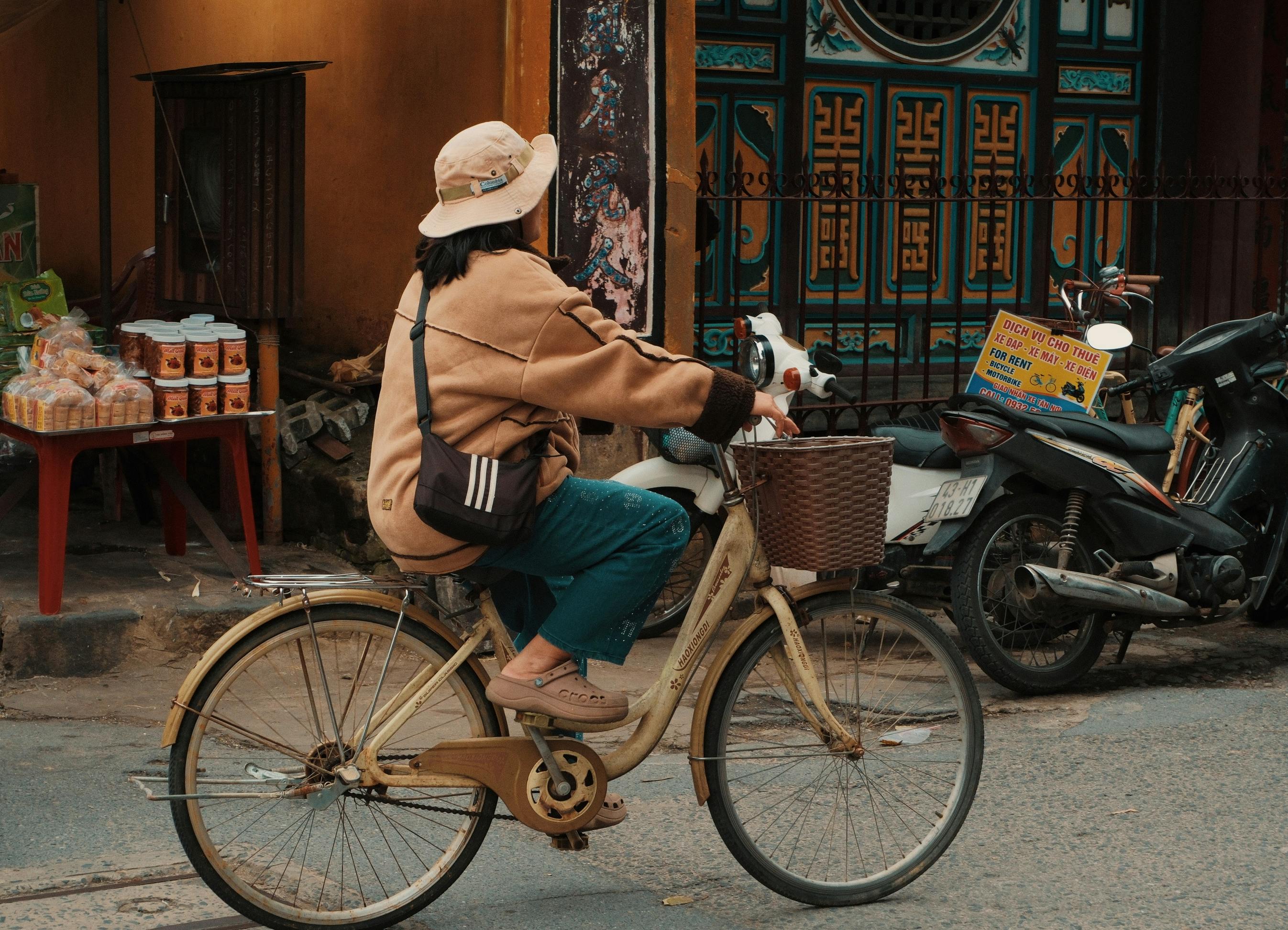 gratis Een vrouw met een hoed fietst door een straat in Đà Nẵng, Vietnam, met een kleurrijke achtergrond. Stockfoto