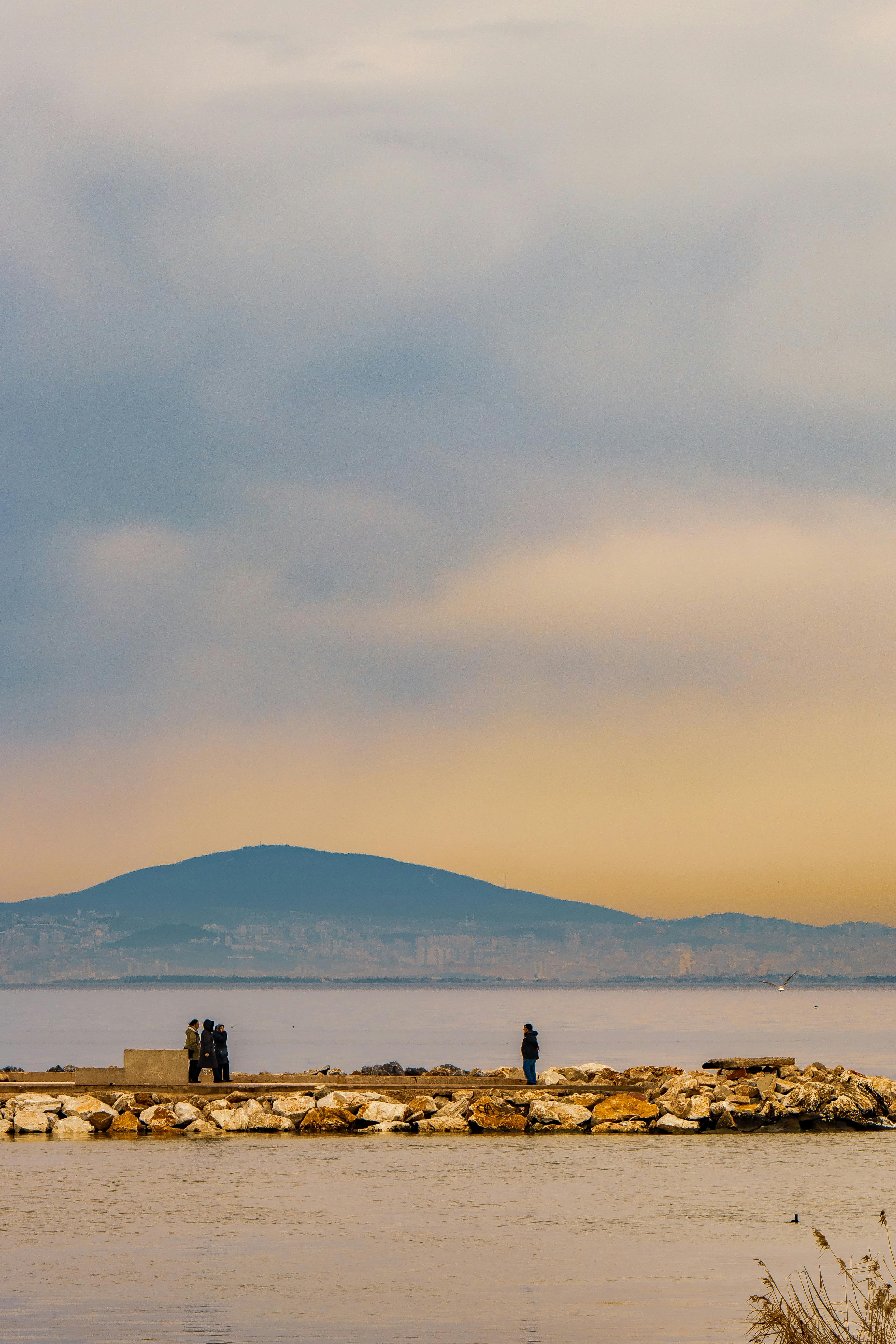Miễn phí Một khung cảnh ven biển thanh bình với những người đi dạo trên lối đi bộ và những ngọn núi xa xa dưới bầu trời màu pastel. Ảnh lưu trữ