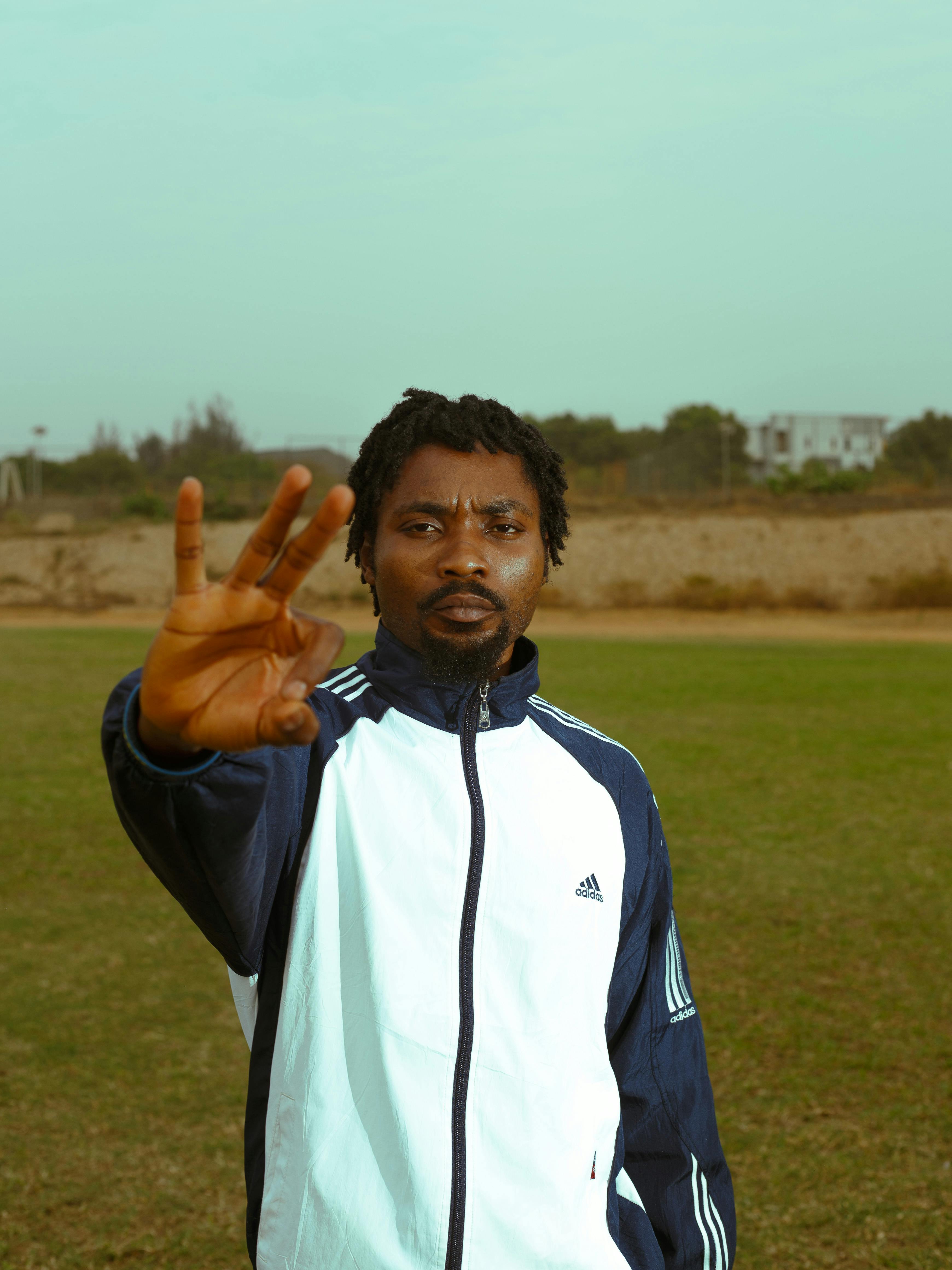 Adult male making a hand gesture in a sports track jacket on a grassy field.