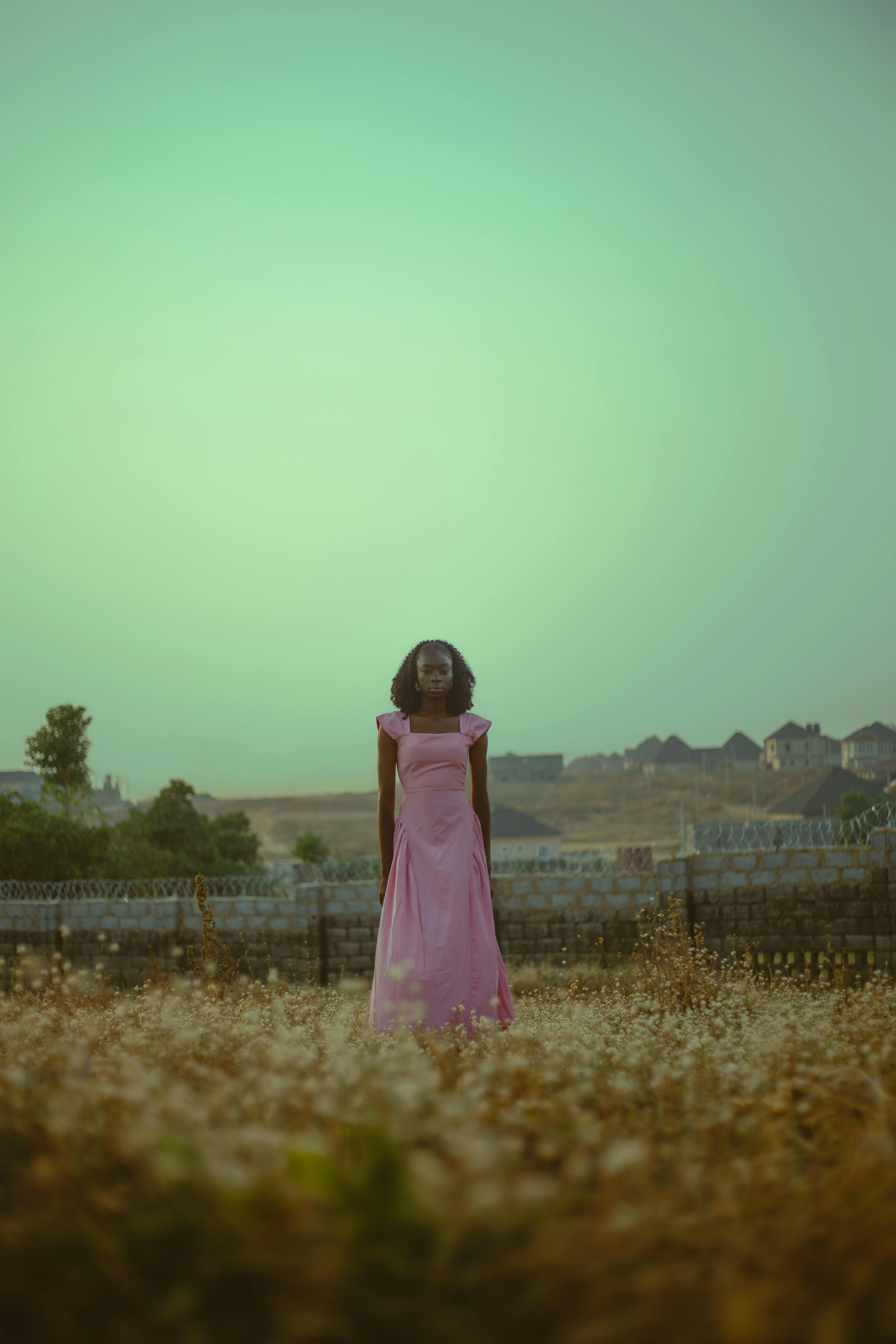 Gratuit O femeie într-o rochie roz, lejeră, stă singură pe un câmp acoperit cu iarbă, emanând calm și liniște. Fotografie de stoc