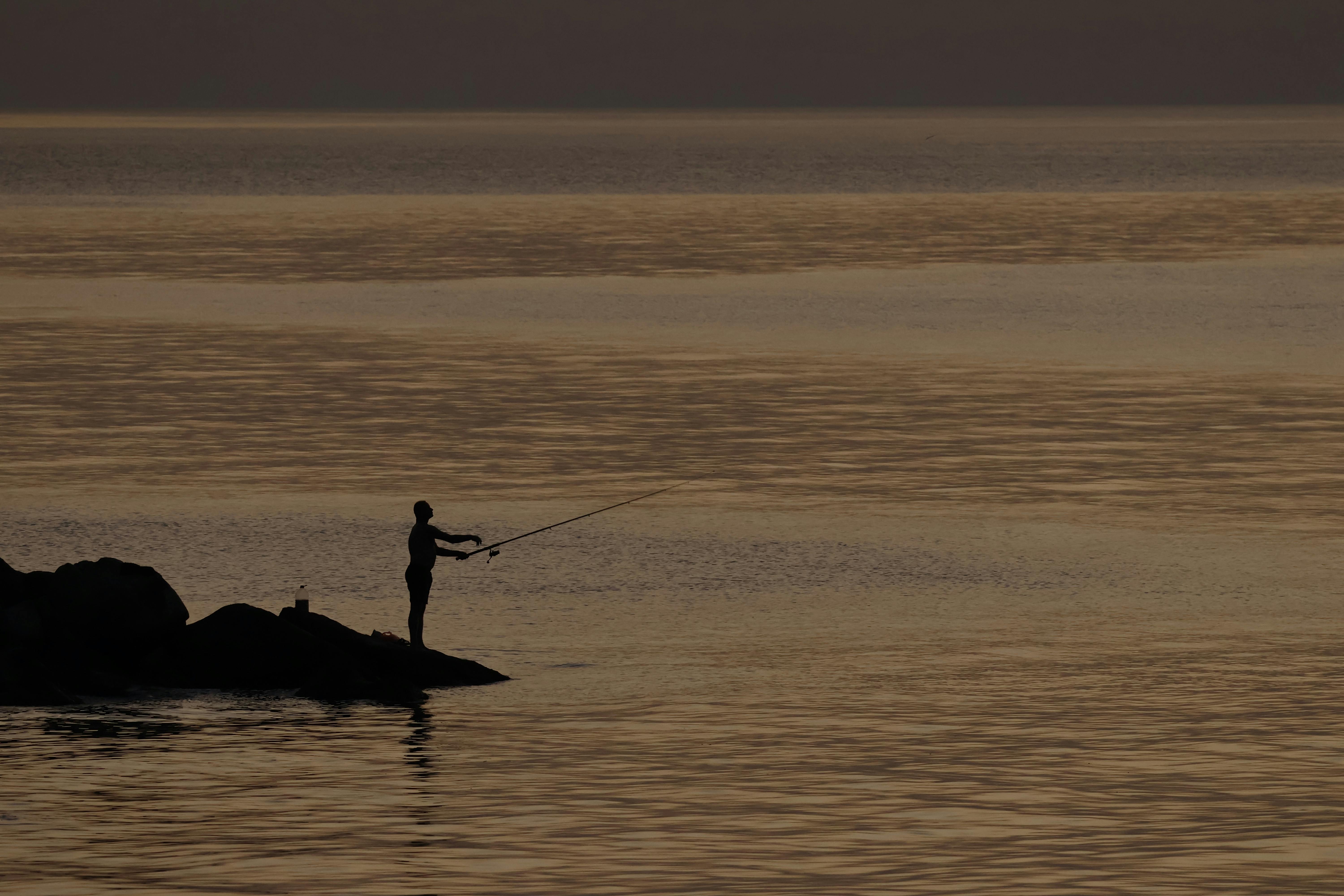 Безкоштовне Самотній рибалка закидає свою волосінь зі скелястого берега в спокійне море на заході сонця. стокове фото