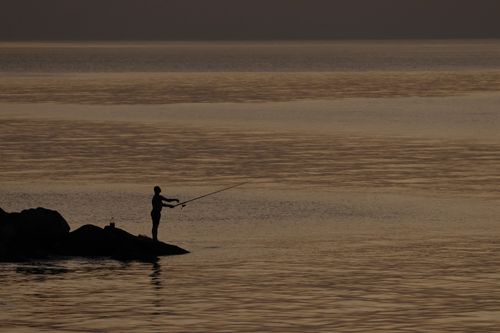Free A lone fisherman casts his line from rocky shore into a serene sea at sunset. Stock Photo