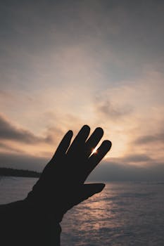 A silhouetted hand reaches towards a beautifully muted sunset sky over water.