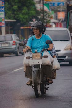 A motorcyclist navigates a busy urban street in rainy weather, carrying grocery bags.