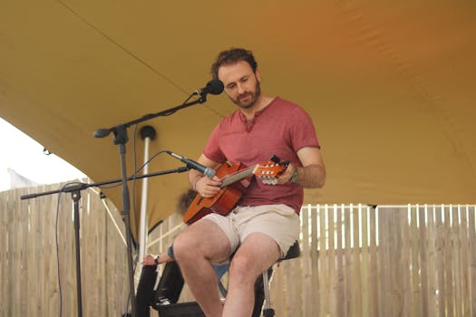 Man playing acoustic guitar on stage during outdoor performance, wearing casual attire.
