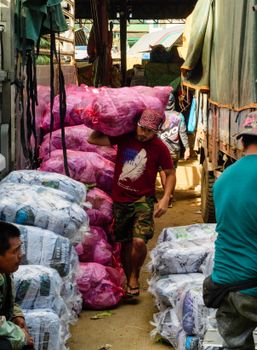 A man carries a large pink sack in a busy outdoor market filled with sacks and goods.