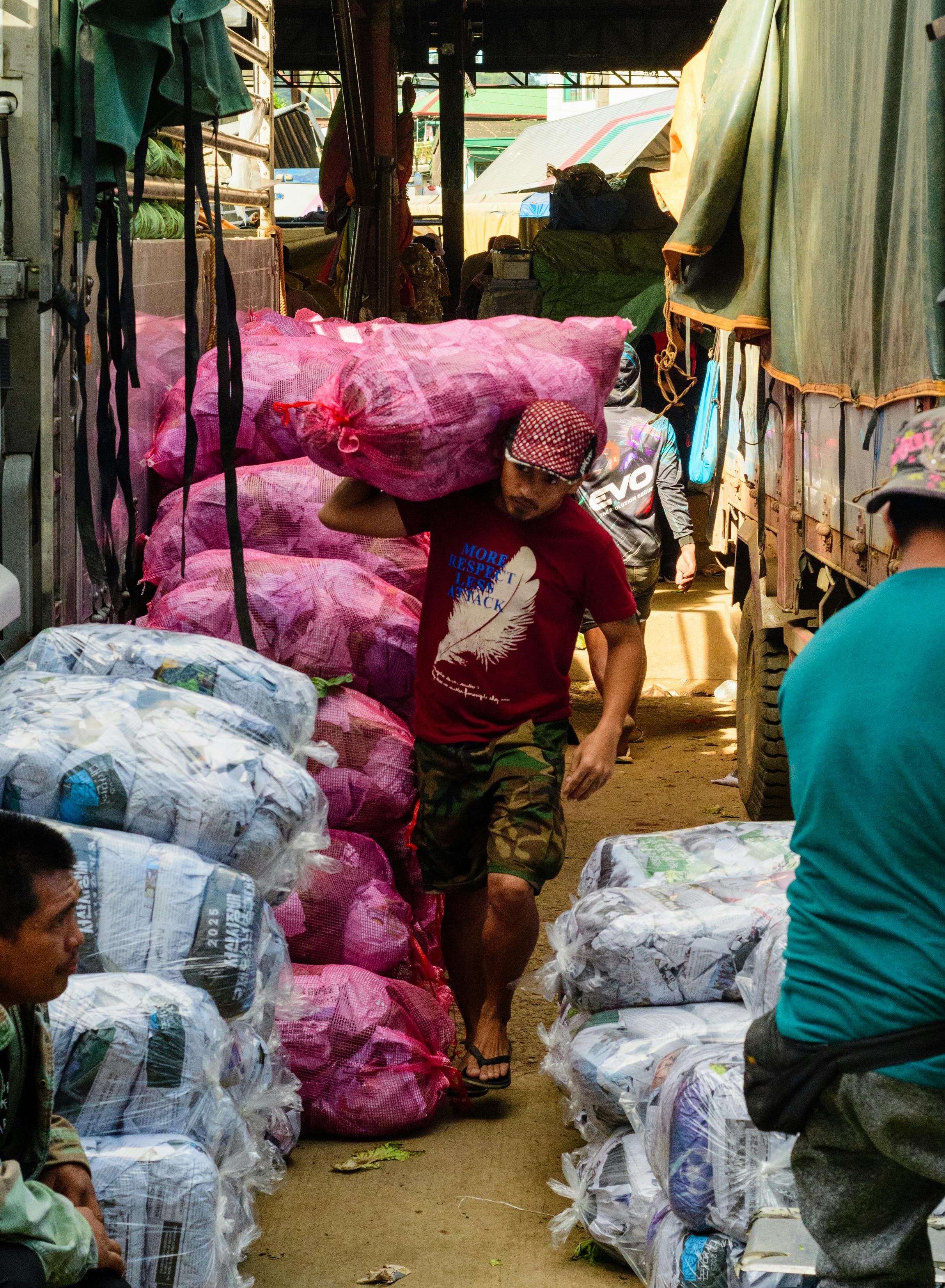 Gratis Un hombre lleva un gran saco rosa en un concurrido mercado al aire libre lleno de sacos y mercancías. Foto de stock