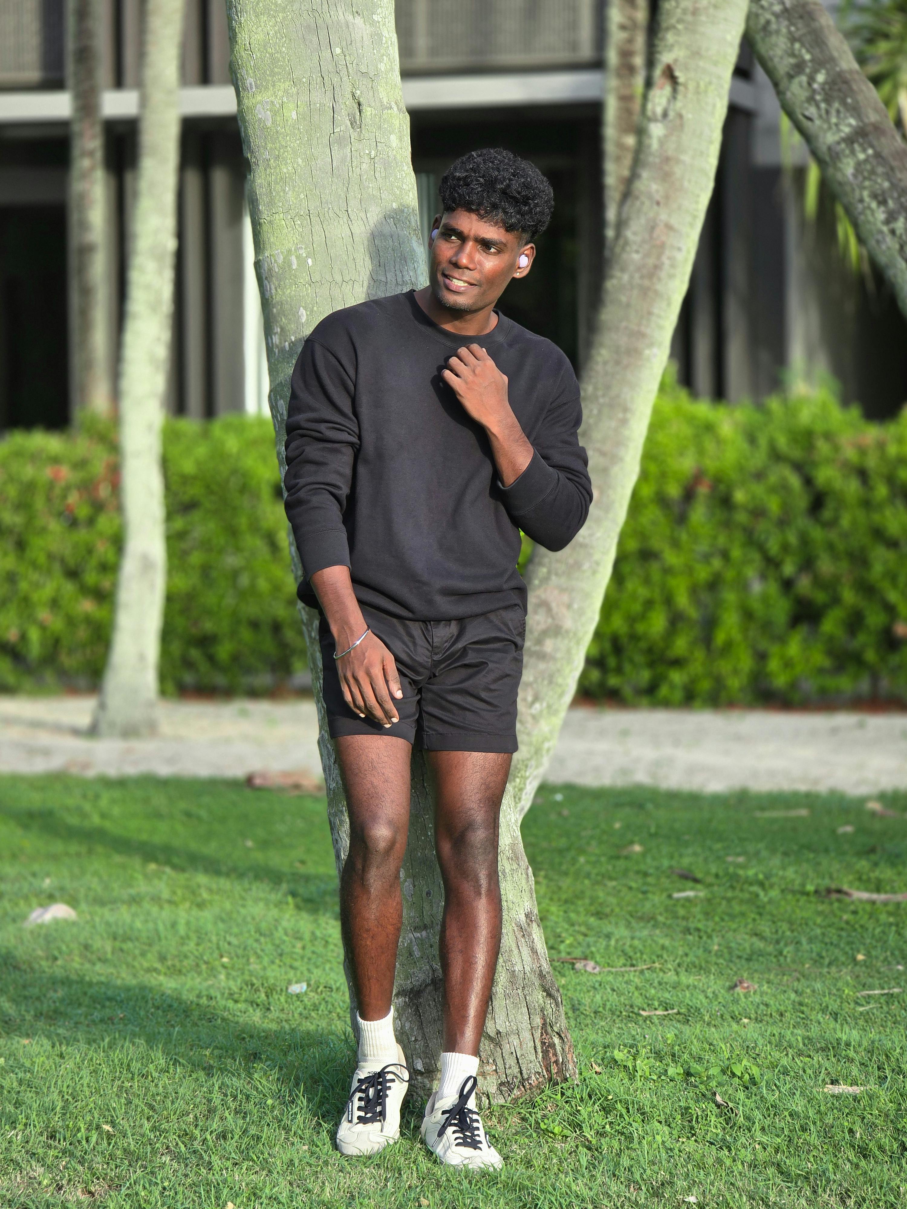 bezplatná Mladý muž v černém oblečení pro volný čas stojící u stromů ve slunném parku. Základová fotografie