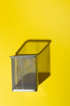 A wire mesh pen holder casting a shadow on a bright yellow background, creating a minimalist effect.