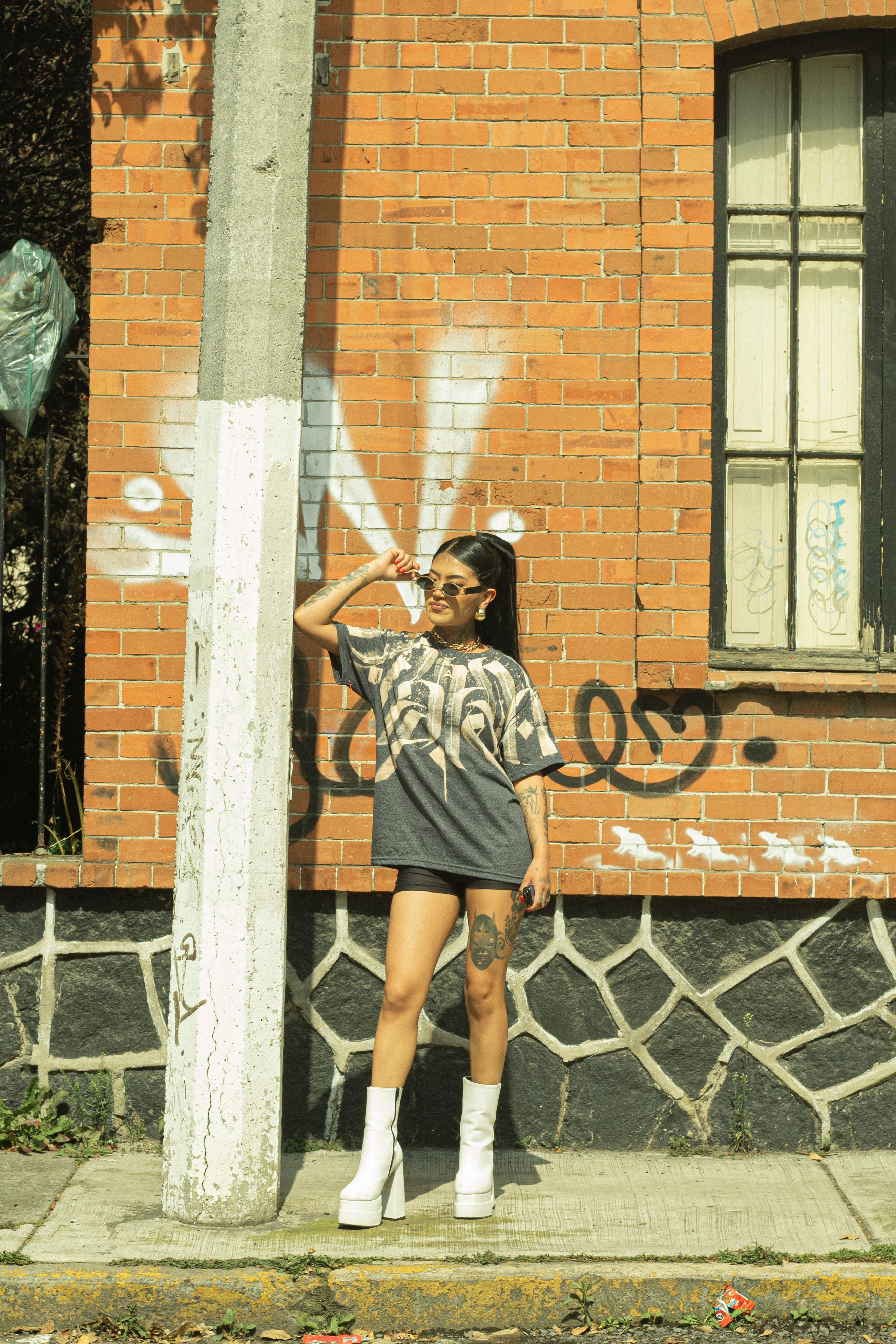 Mulher estilosa de óculos escuros e botas posando ao lado de uma parede de tijolos coberta de grafite à luz do dia.