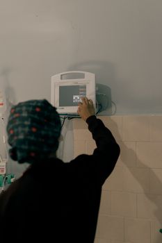 A healthcare worker with a head covering using medical monitoring equipment indoors.