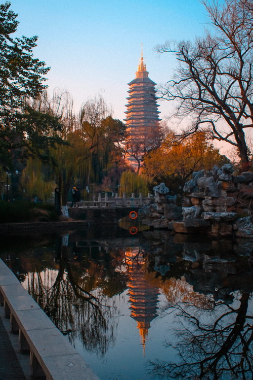 Free A tranquil scene with a pagoda reflected in a park pond at sunset, surrounded by autumn trees. Stock Photo