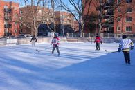 Adults playing ice hockey on an outdoor rink in a city during winter.