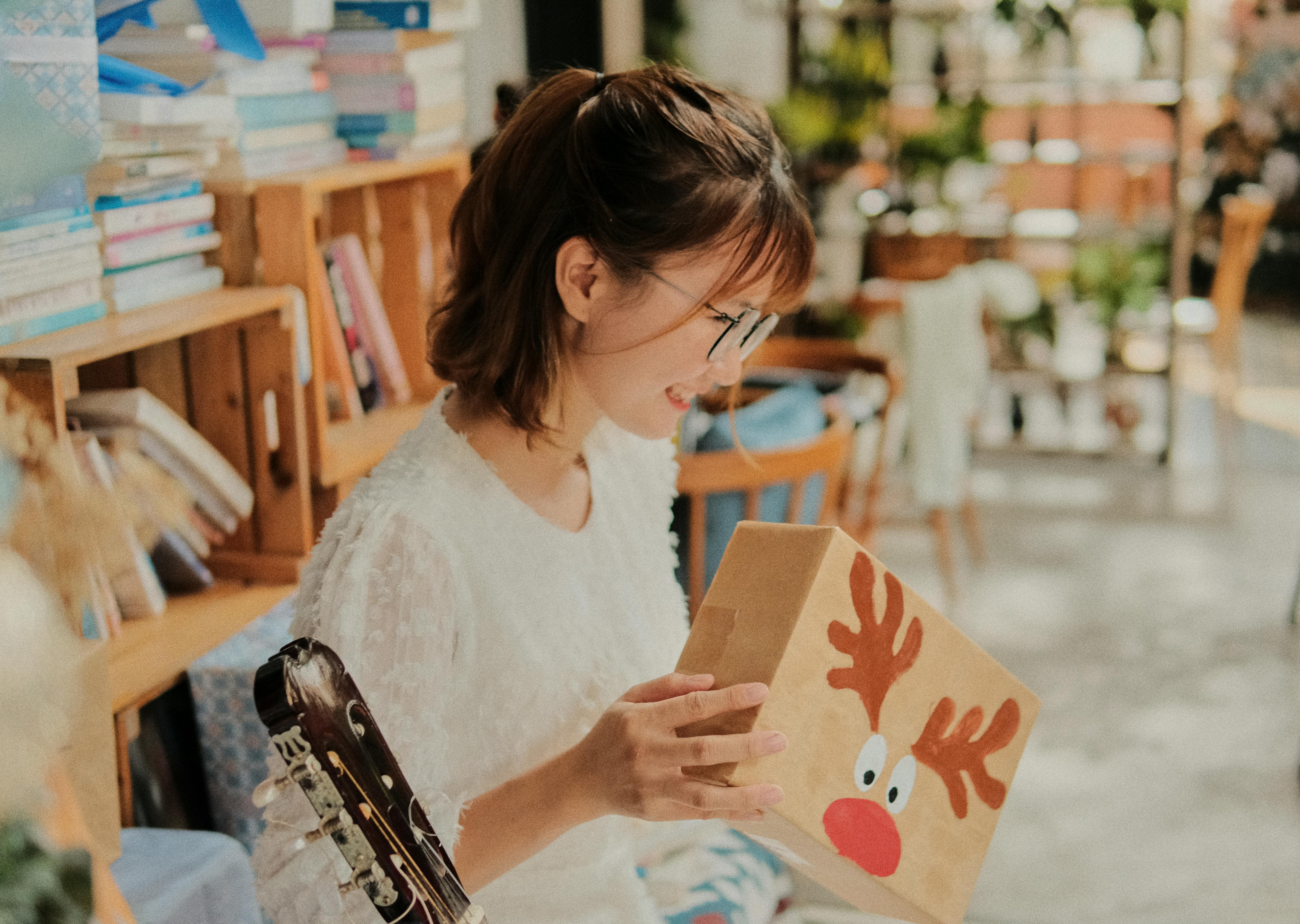 Gratis Una gioiosa donna vietnamita in abito bianco scarta un regalo di Natale a tema renna all'interno di un'accogliente biblioteca. Foto a disposizione