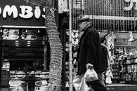 A black and white photo of an elderly man walking in an urban street setting, holding grocery bags.
