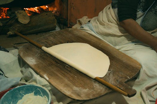 A woman prepares traditional Turkish flatbread by a fire using a rolling pin and wooden board.