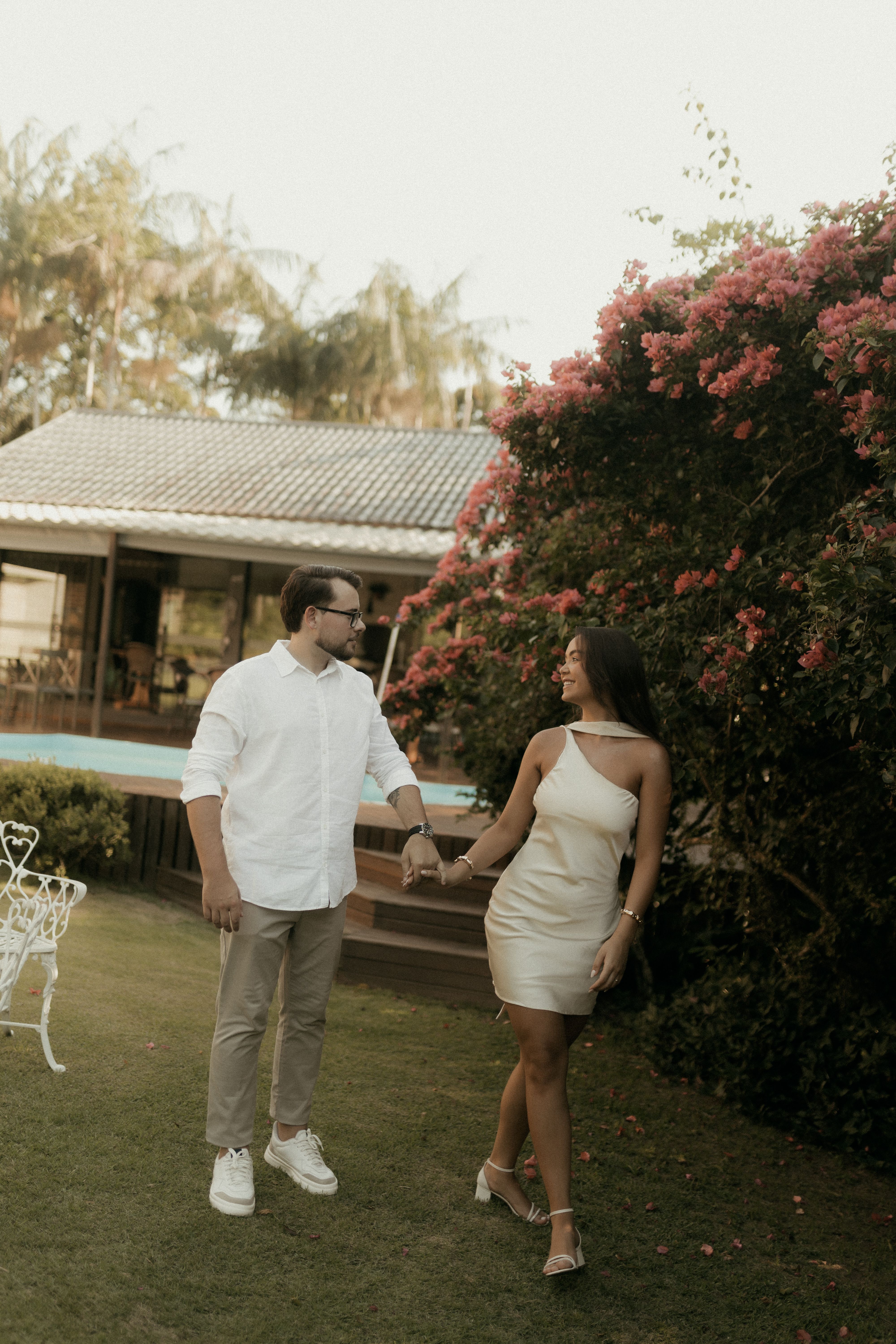 A couple enjoys a romantic walk in a lush garden, surrounded by vibrant flora and cozy architecture.