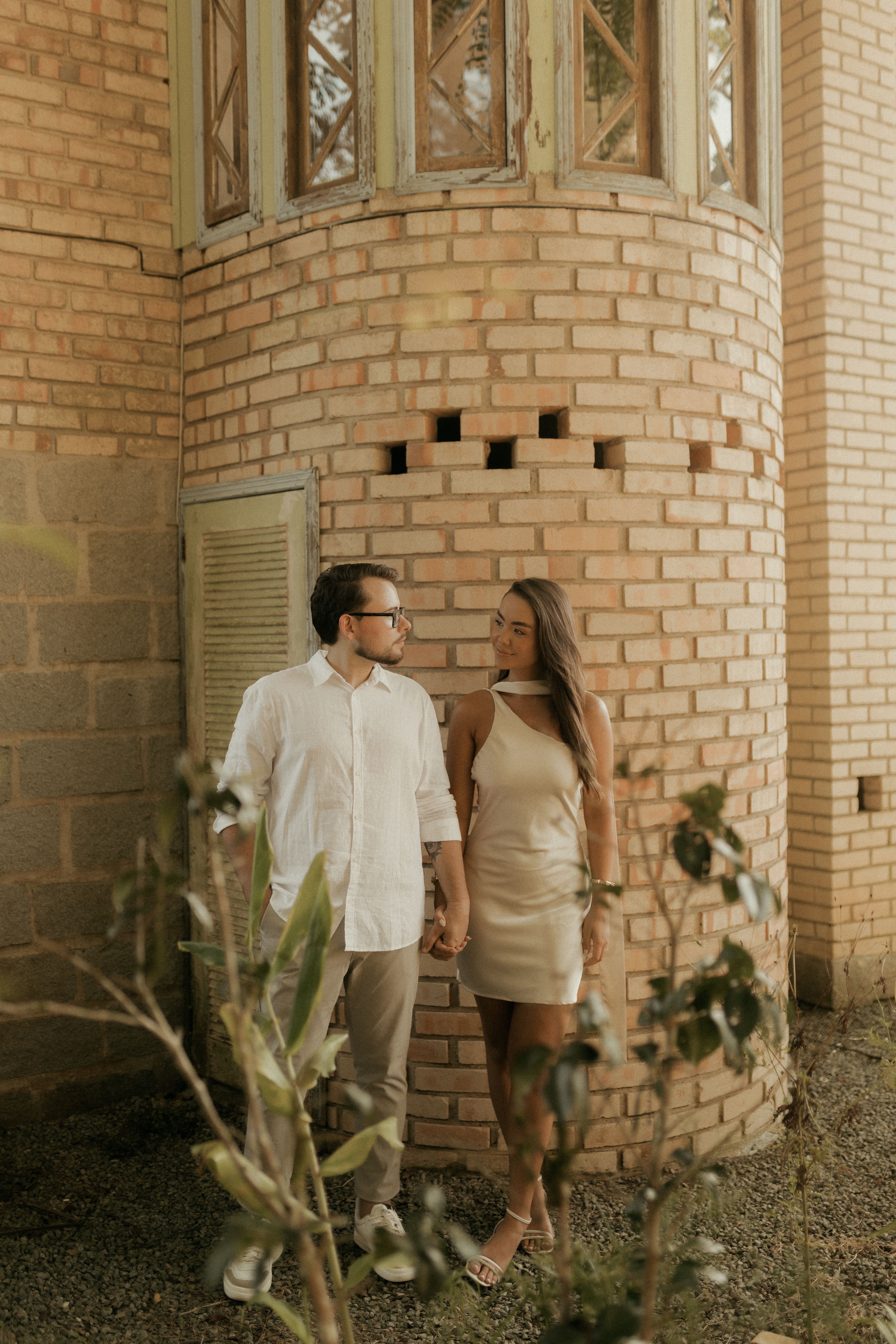 A couple romantically holding hands by a vintage brick building, surrounded by lush foliage.
