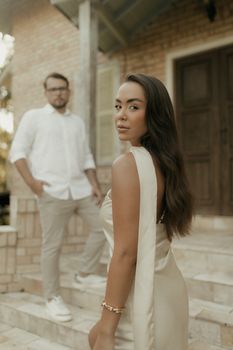 A stylish couple poses romantically on steps of a rustic house in warm daylight.