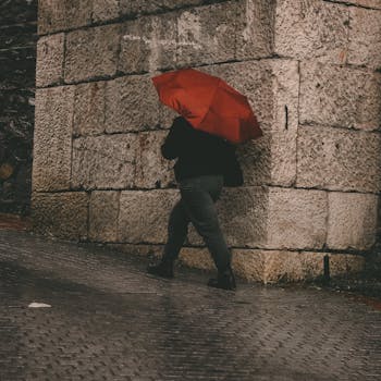 Silhouetted figure walking with a vibrant red umbrella against a rugged stone wall on a rainy day.