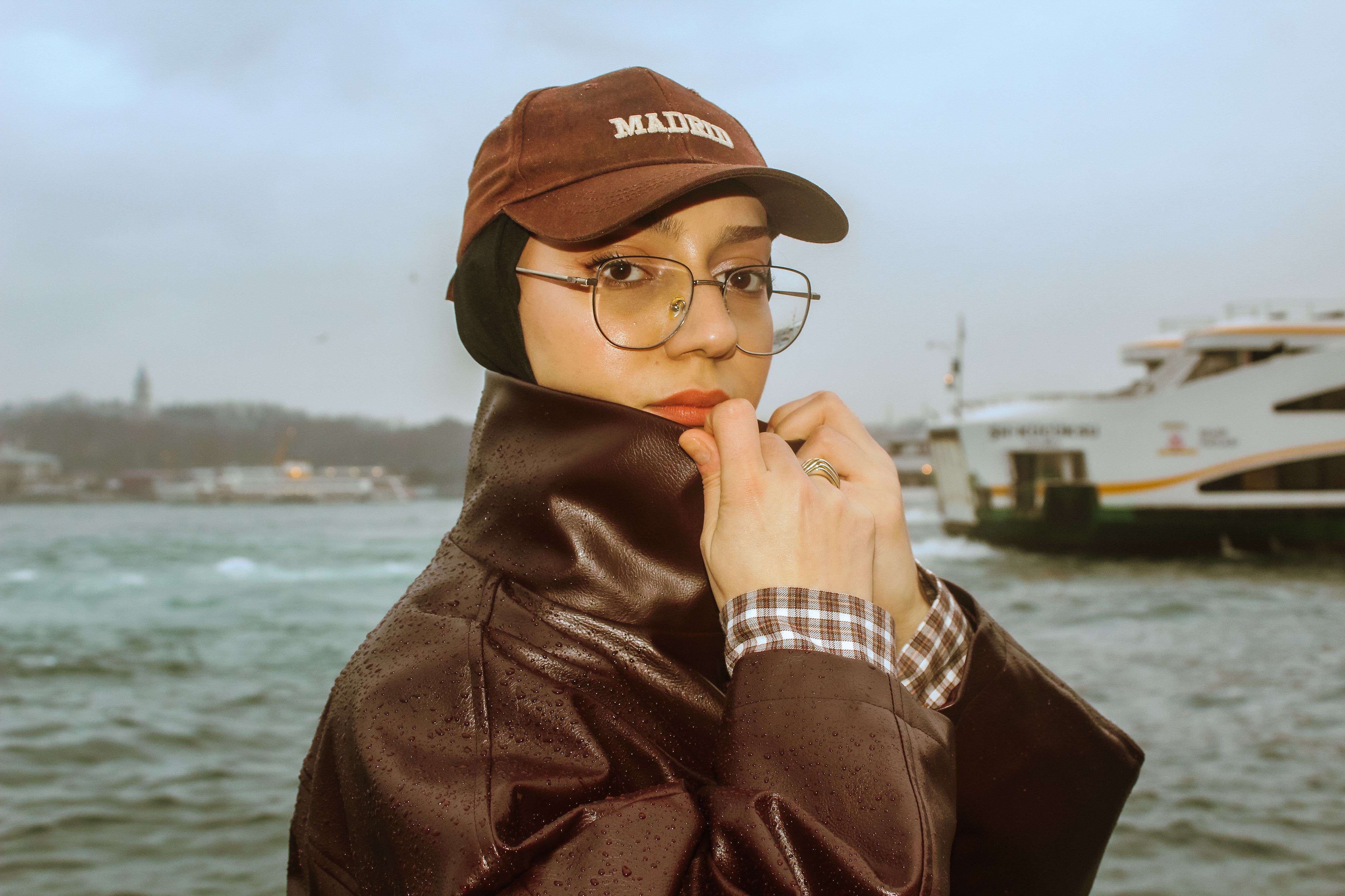A woman in a raincoat and cap stands by a waterfront, with boats visible in the background.