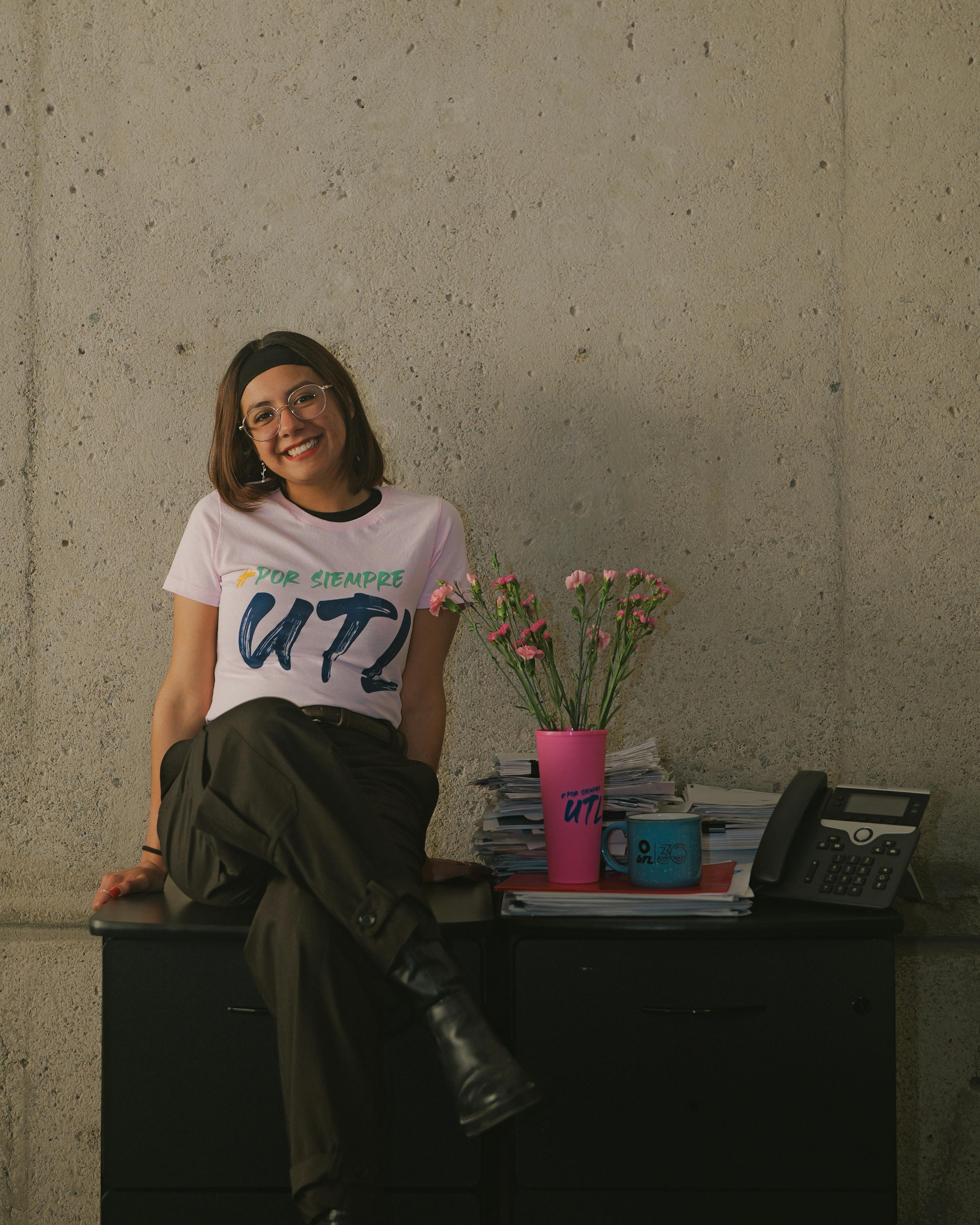 Gratis Donna felice seduta alla scrivania nell'ufficio dell'università, circondata da fiori e in un'atmosfera allegra. Foto a disposizione