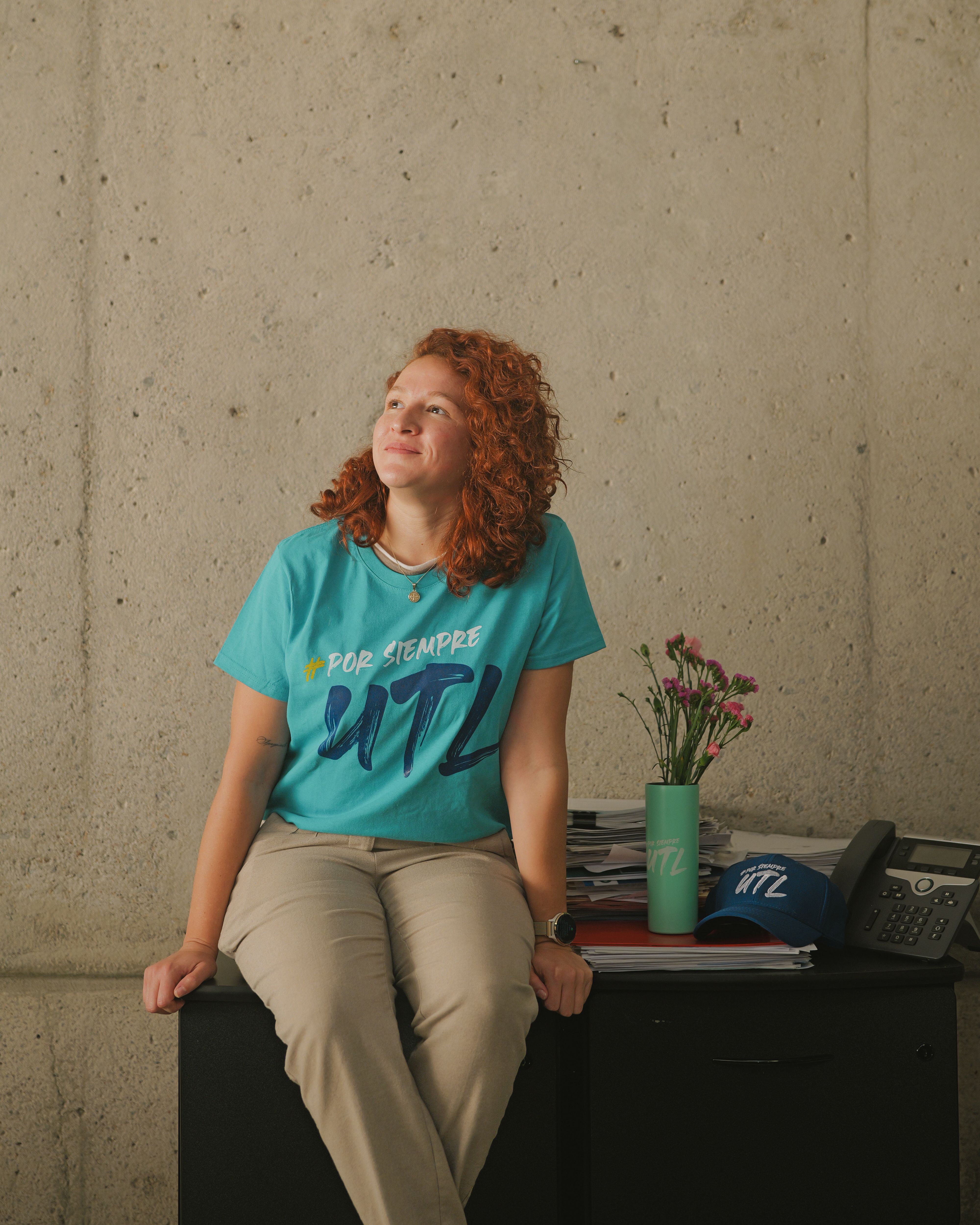 Red-haired woman sitting on a desk in a relaxed pose, wearing casual clothes indoors.