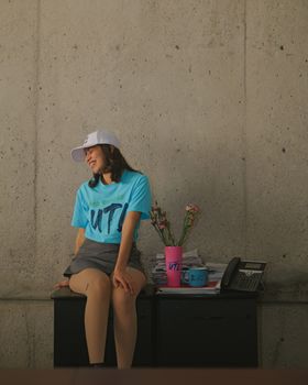 Young woman sitting on desk indoors, dressed casually, with a phone and mugs around.