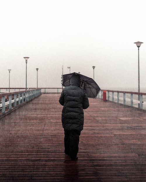 Person Wearing Black Jacket and Black Pants Holding Umbrella Walking on Boardwalk
