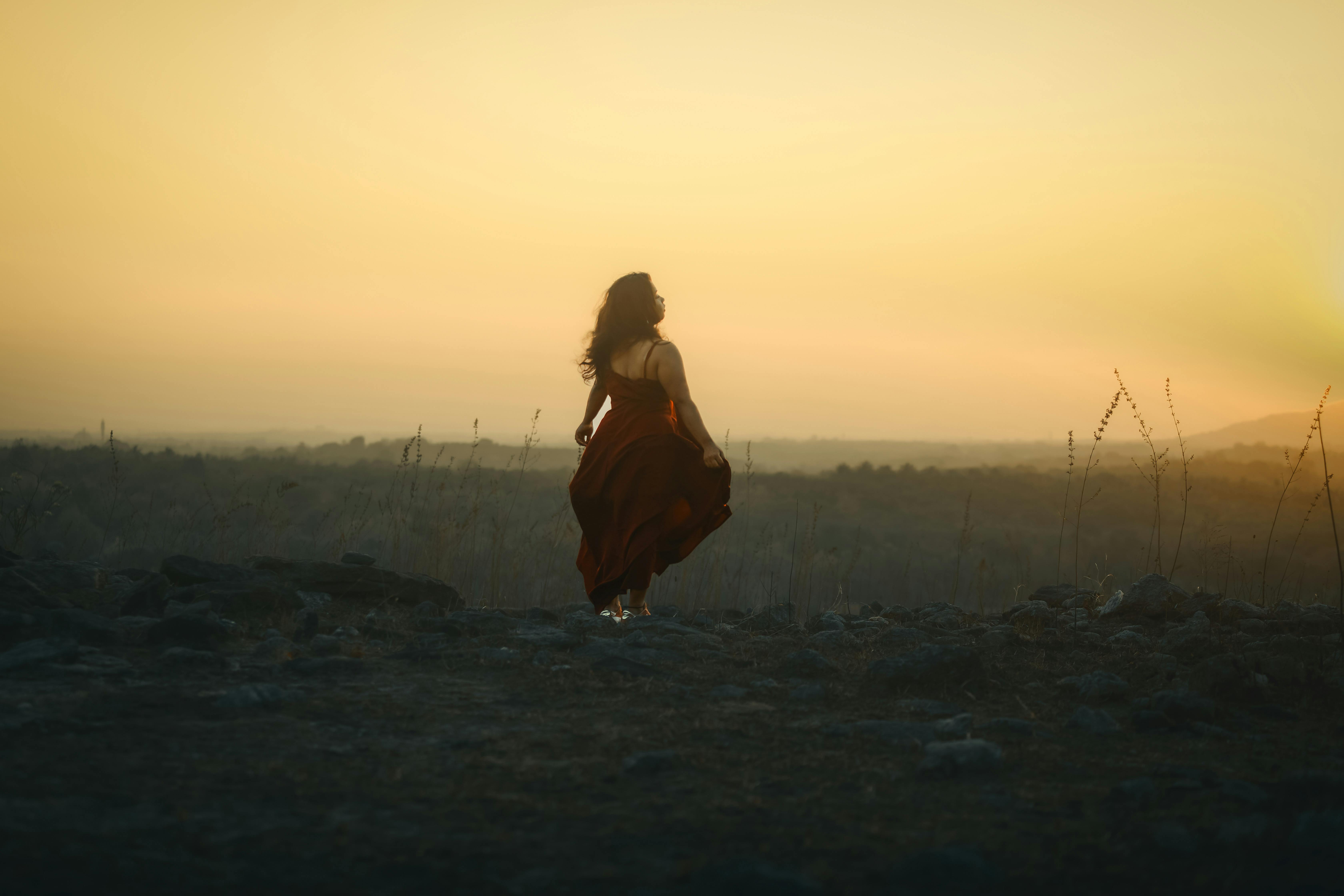 gratis Een vrouw in een zwierige rode jurk staat in een open veld tijdens een warme zonsondergang, wat een serene en dromerige scène oplevert. Stockfoto