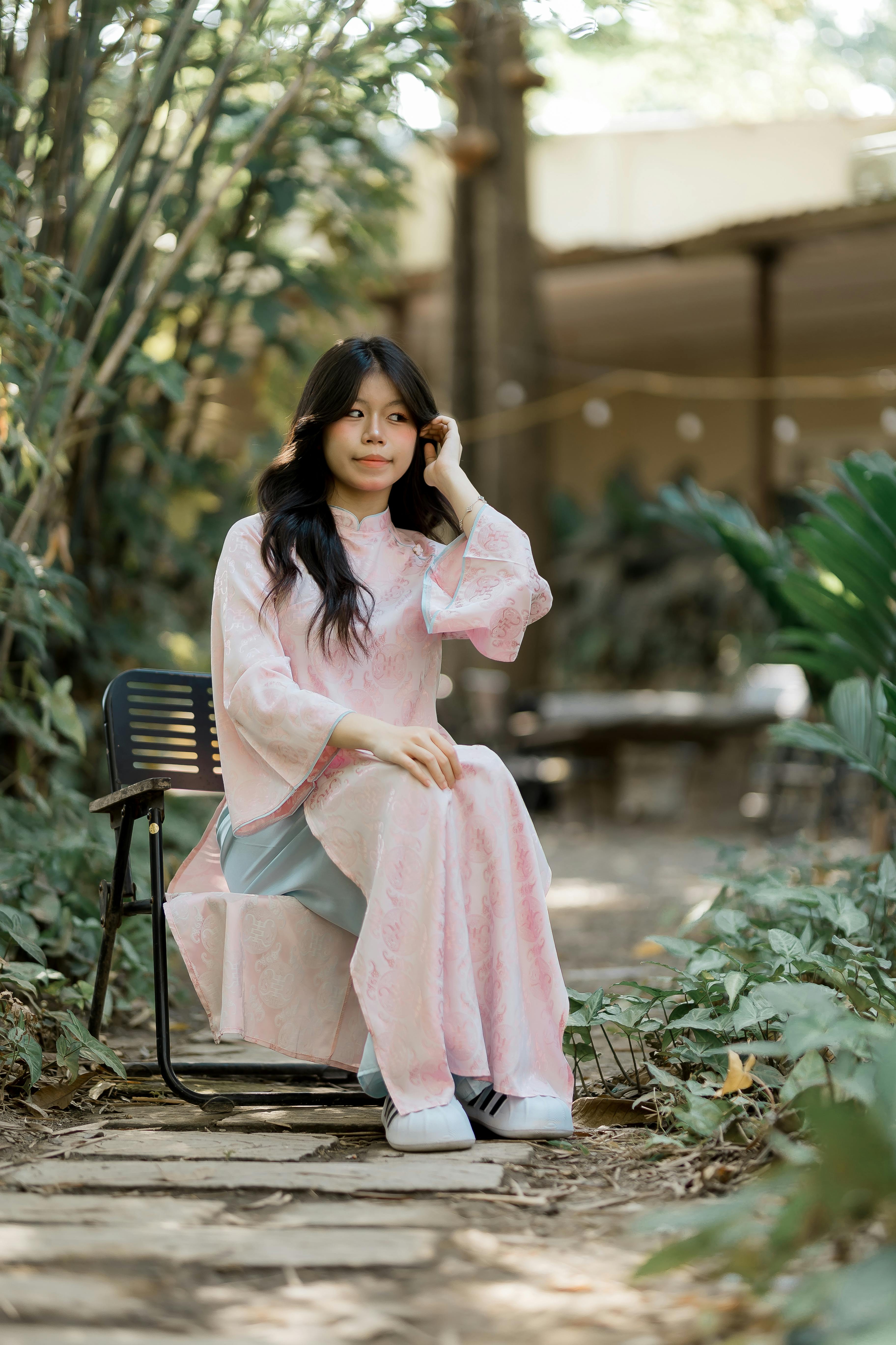 ฟรี หญิงสาวในชุดเดรสสีชมพูนั่งอยู่บนเก้าอี้กลางแจ้ง ท่ามกลางต้นไม้ใบไม้ คลังภาพถ่าย