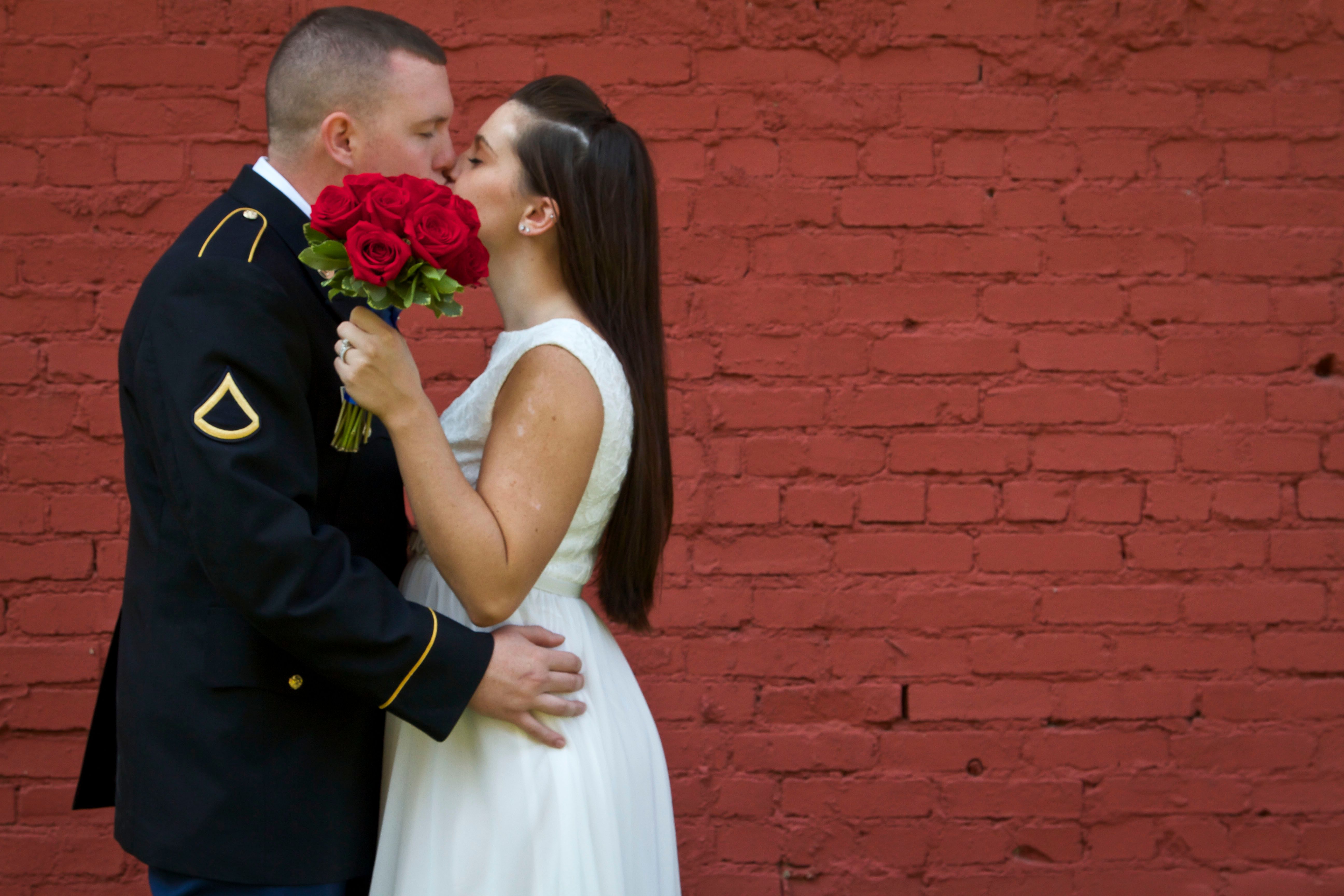A military groom and bride share a romantic kiss against a red brick wall, symbolizing love and commitment.