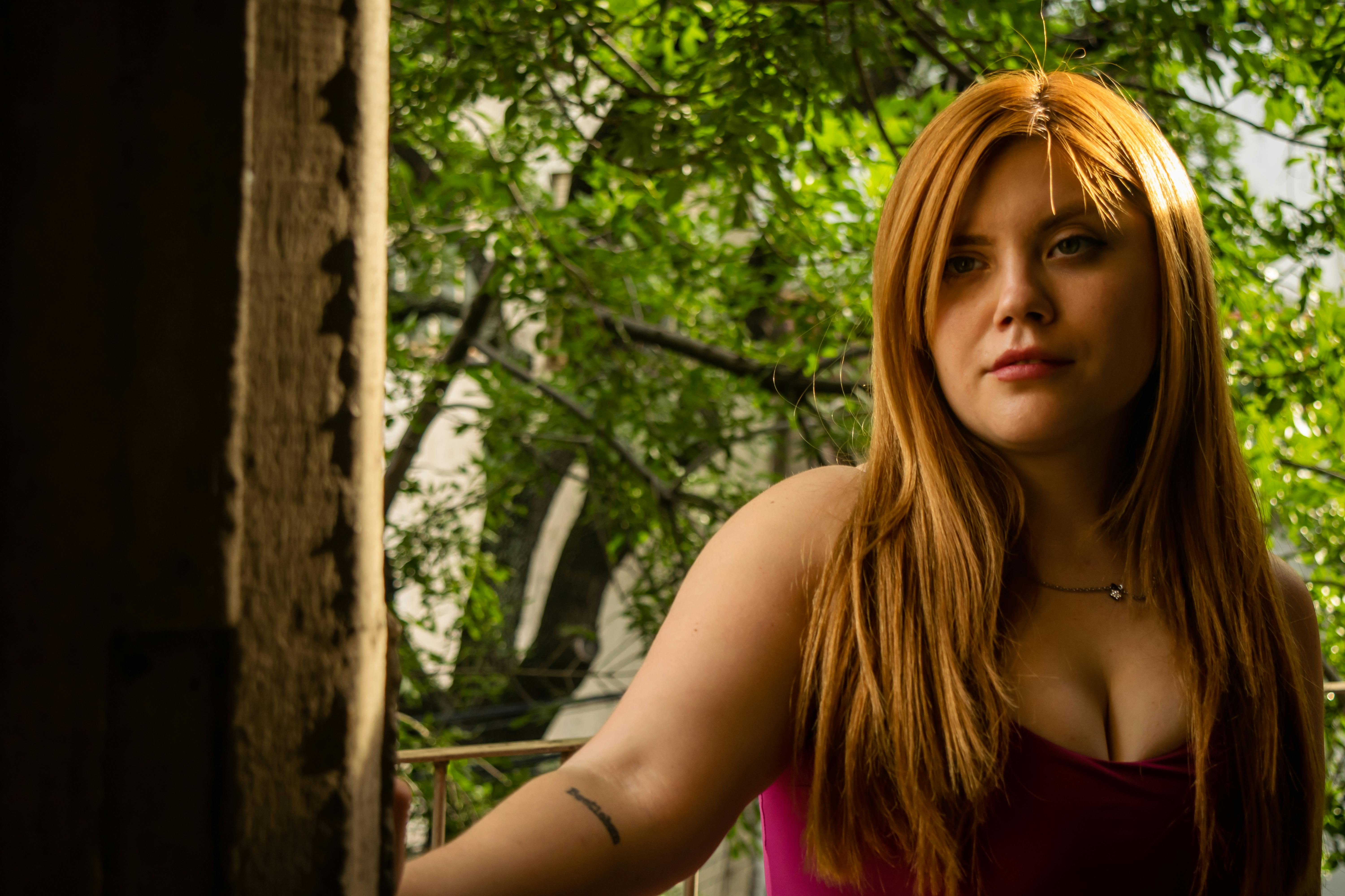 Gratis Ritratto di una giovane donna con i capelli rossi in piedi vicino a una finestra, circondata dal verde. Foto a disposizione