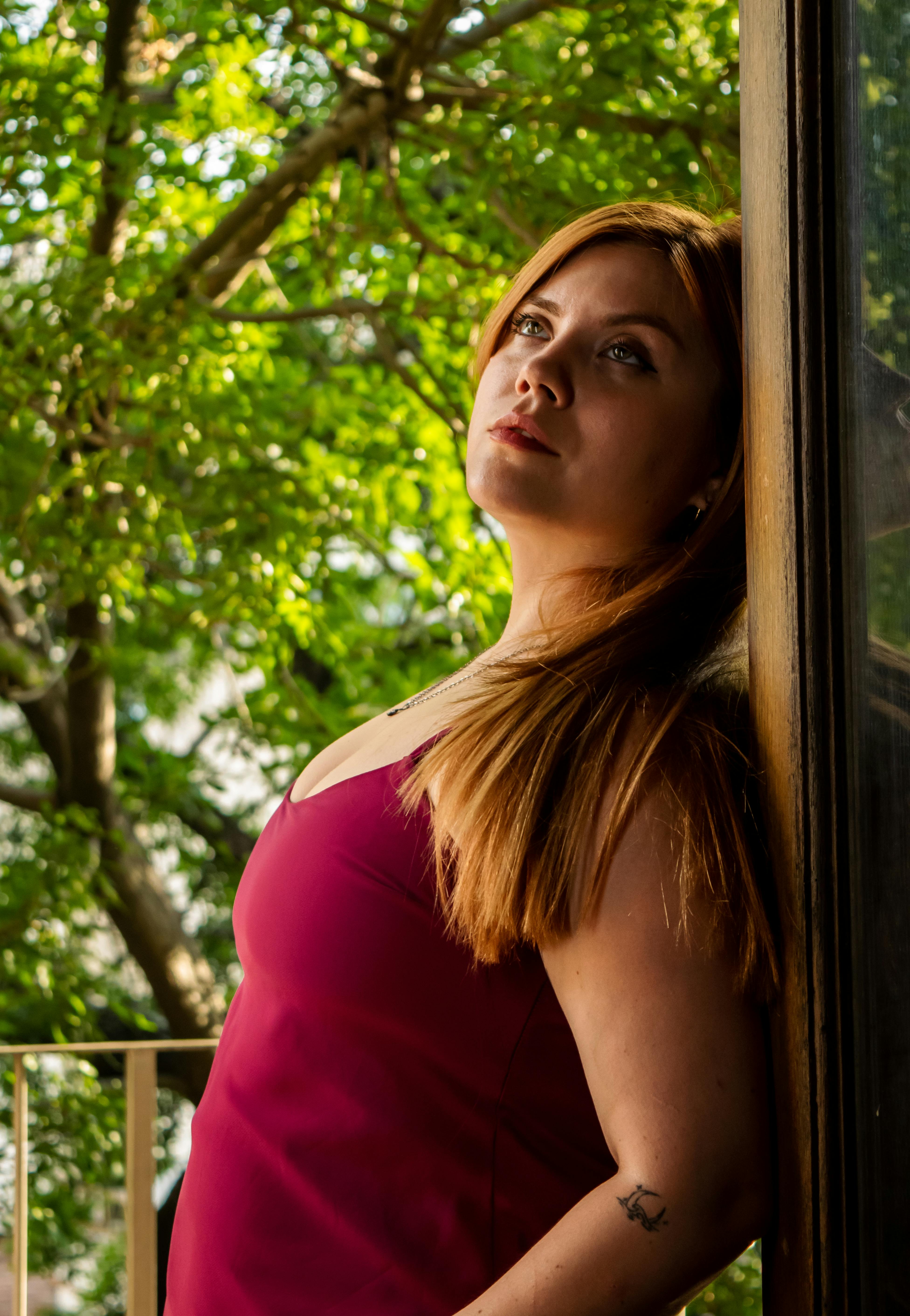Free A thoughtful woman in a red dress leans against a balcony railing surrounded by vibrant green foliage. Stock Photo