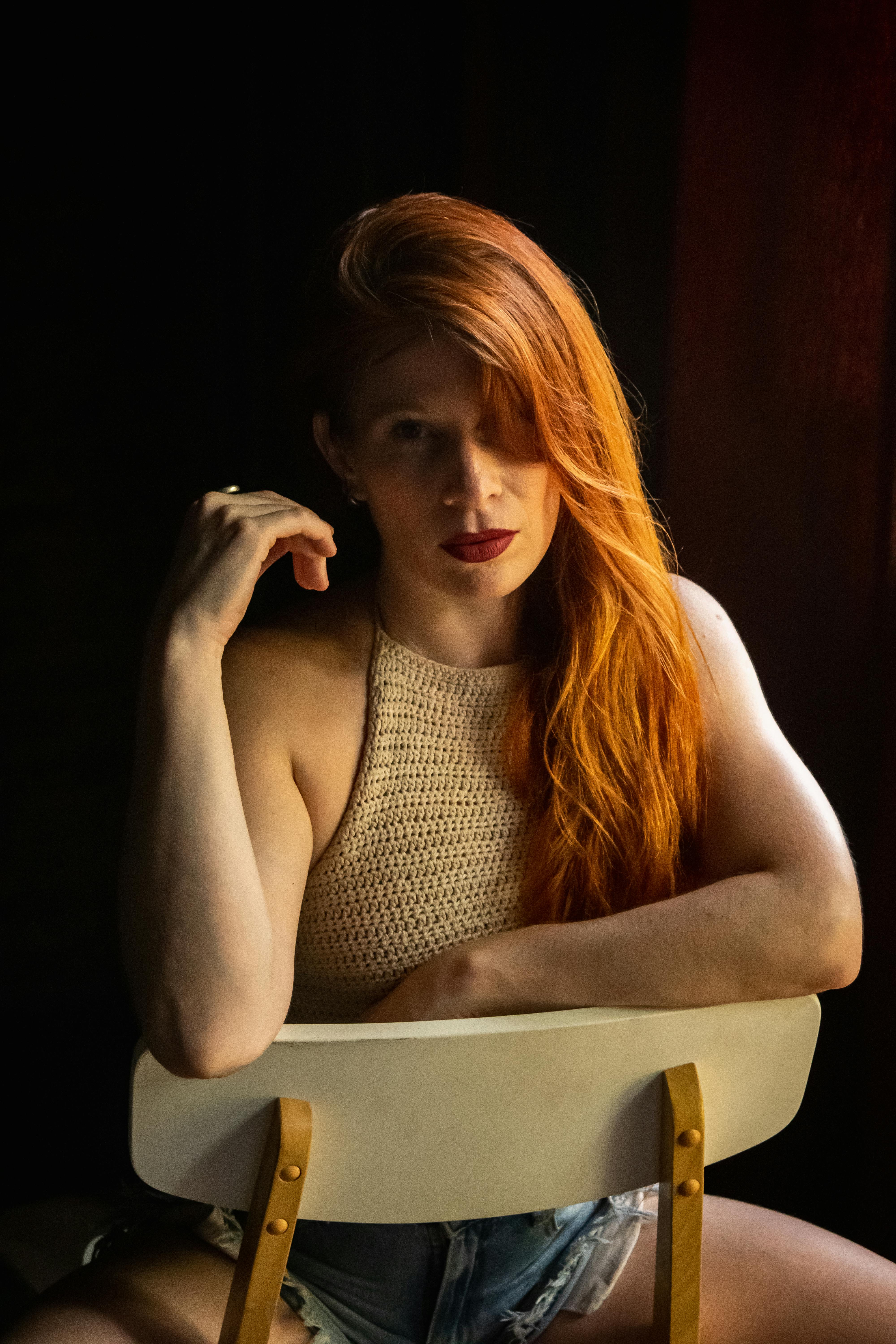 Gratis Affascinante ritratto di una donna dai capelli rossi seduta su una sedia, illuminata dalla luce naturale. Foto a disposizione