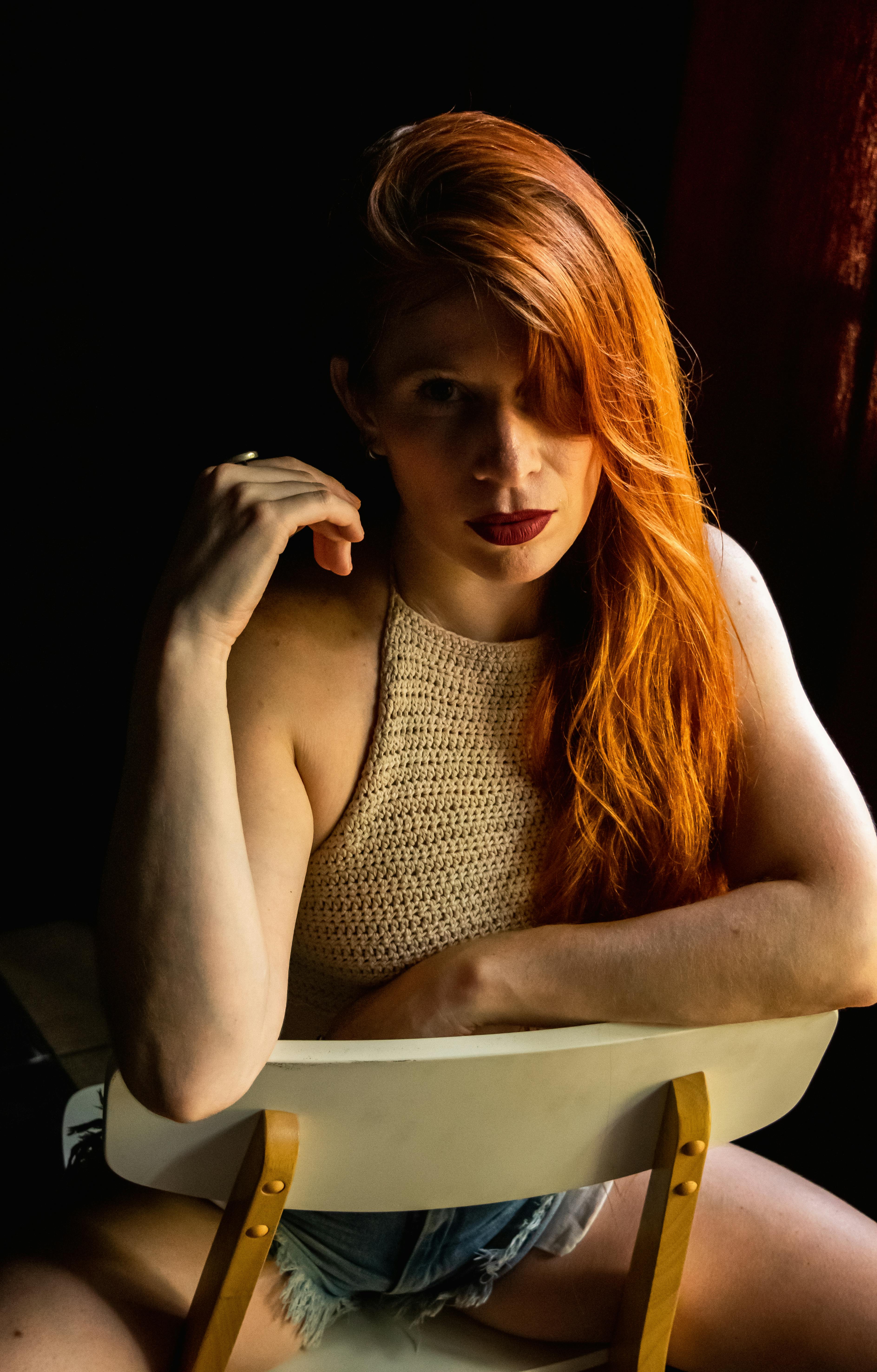 ฟรี ภาพเหมือนศิลปะของหญิงสาวผมแดงนั่งอยู่บนเก้าอี้ในแสงสลัวๆ คลังภาพถ่าย