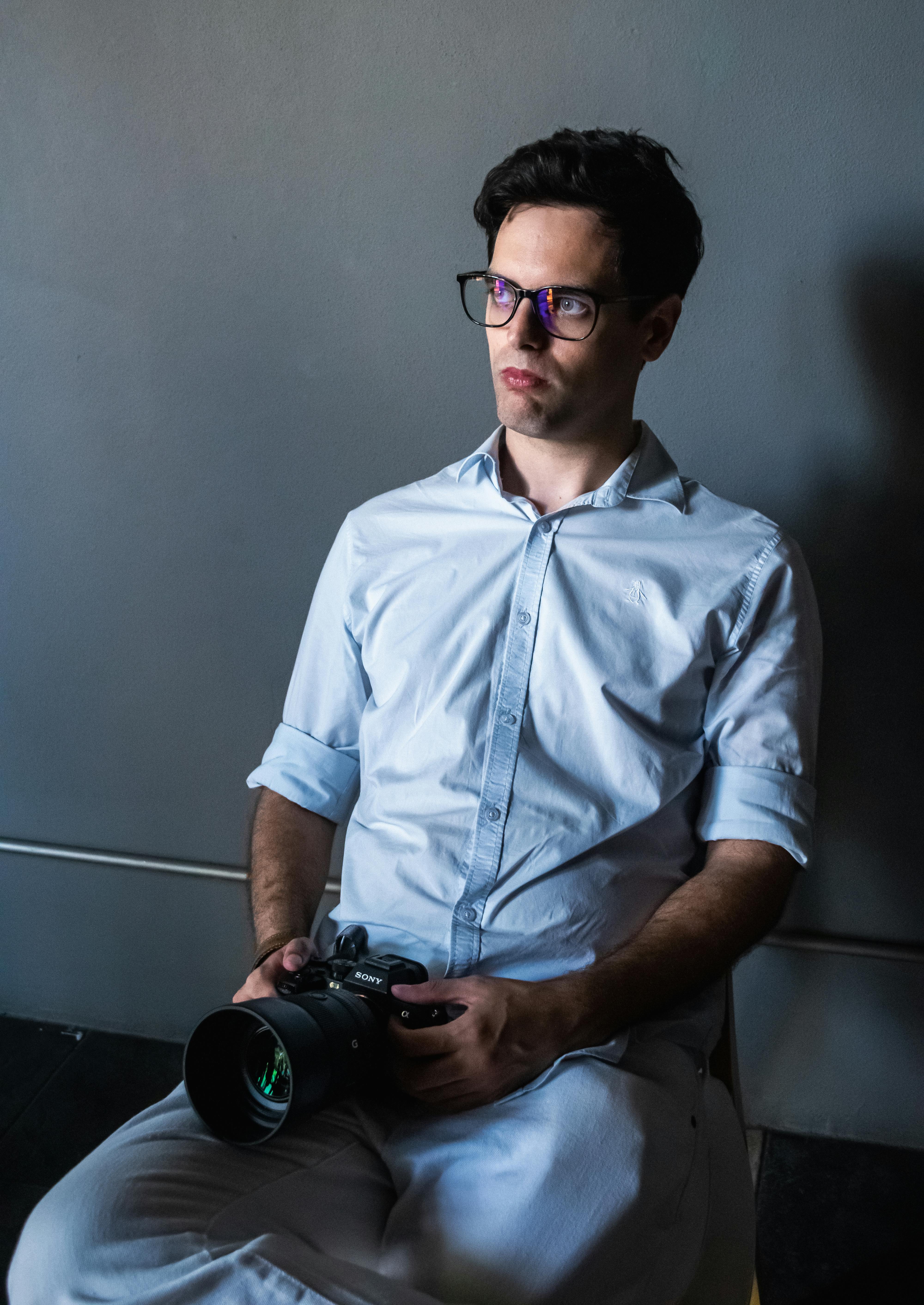 Gratis Un giovane pensieroso che tiene in mano una macchina fotografica in uno studio con luci soffuse. Foto a disposizione