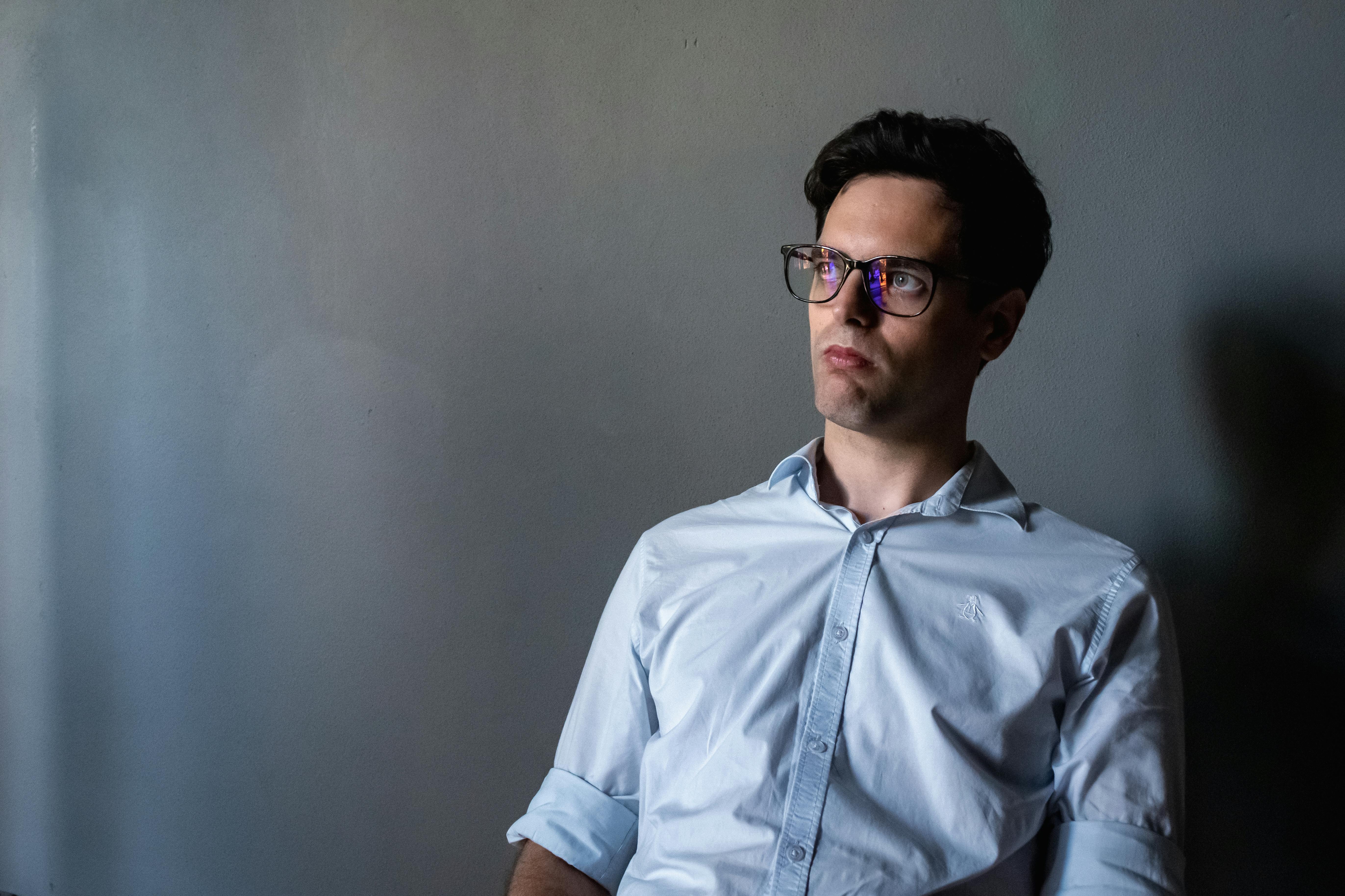 Miễn phí Một người đàn ông trầm tư đeo kính ngồi trong một căn phòng thiếu ánh sáng, mặc áo sơ mi màu xanh nhạt và chăm chú nhìn xung quanh. Ảnh lưu trữ