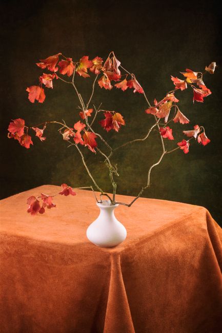 Vibrant autumn leaves arranged in a sleek white vase, set atop a rust-colored tablecloth, evoking a warm fall ambiance.