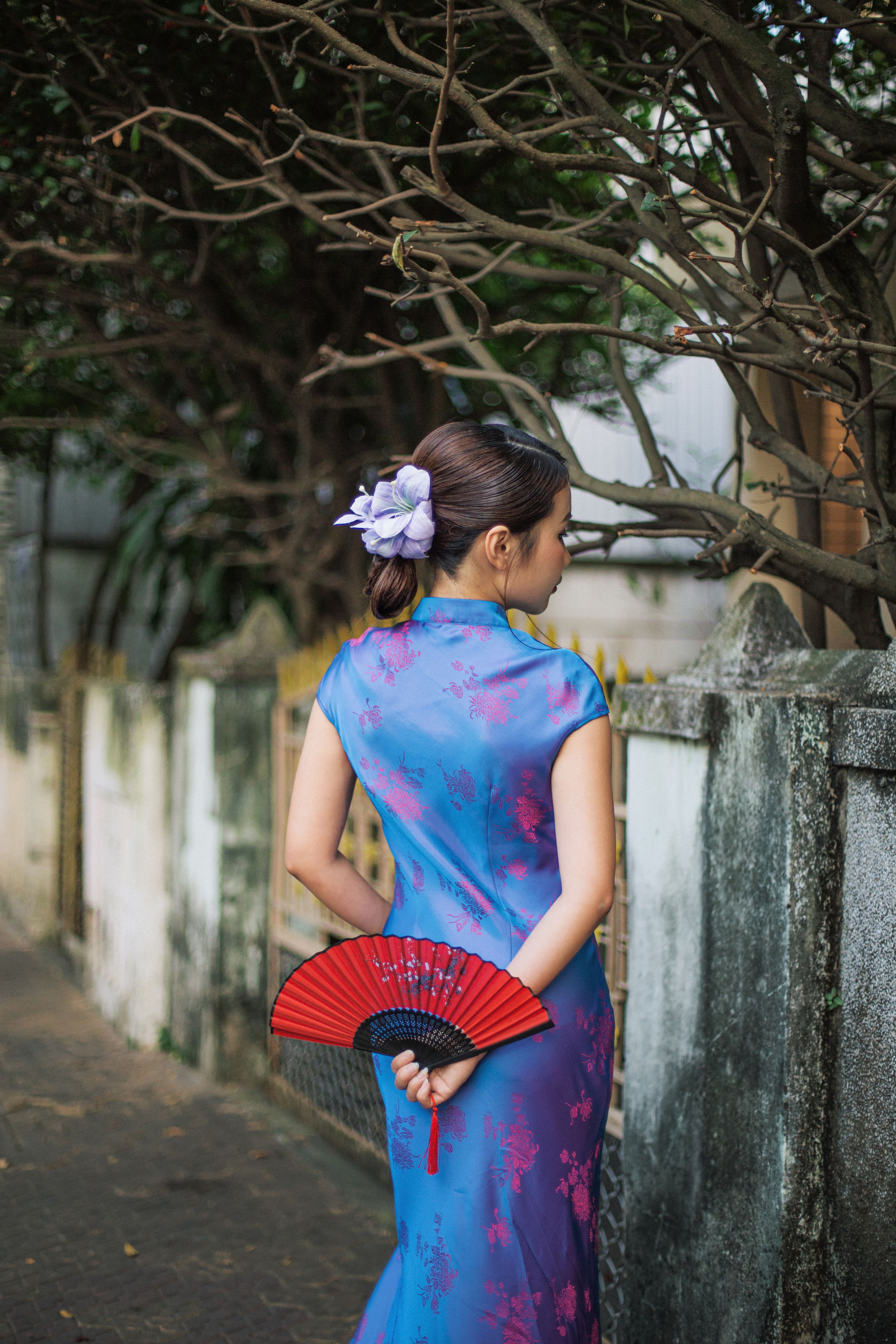 Woman in a blue cheongsam holding a red fan, standing outdoors by a wall.