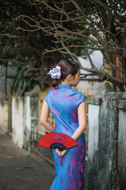 Woman in a blue cheongsam holding a red fan, standing outdoors by a wall.