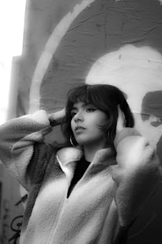 Black and white portrait of a young woman in a fur coat, leaning against a graffiti wall.